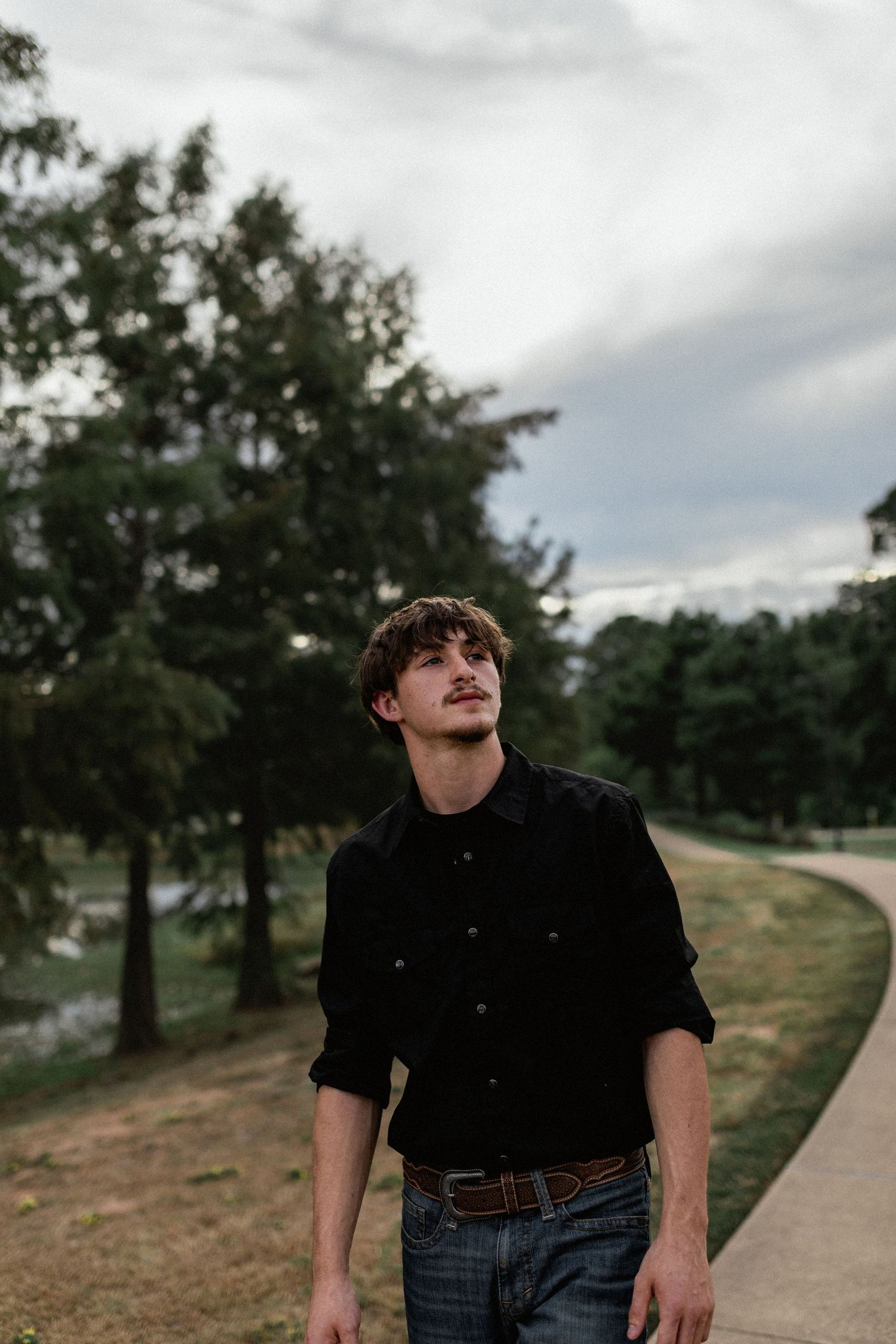 Young man in black shirt and jeans walking along a park path under a cloudy sky.