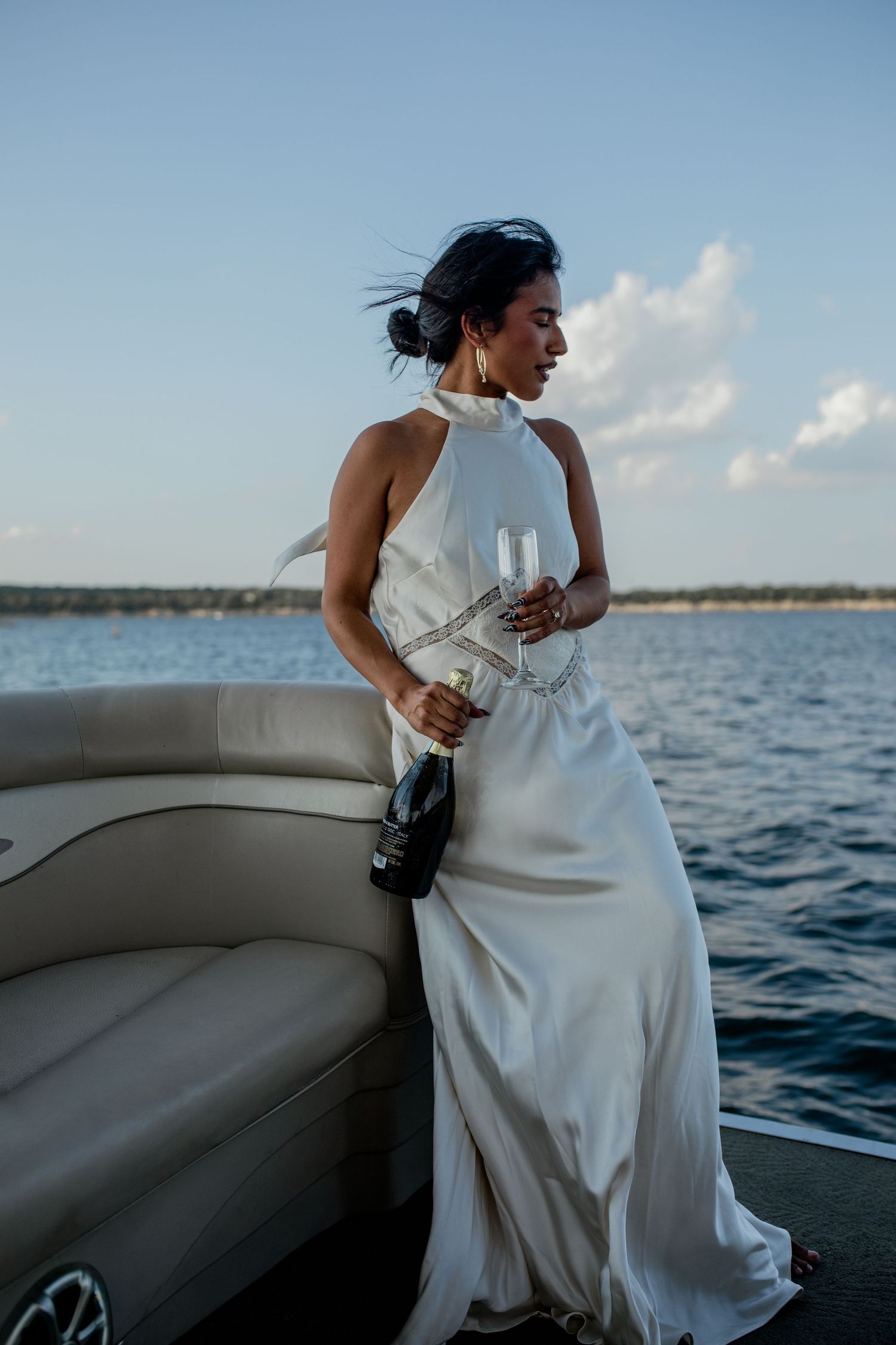 Elegant woman in white halter gown holding champagne bottle and flute on a boat at sunset over calm water.