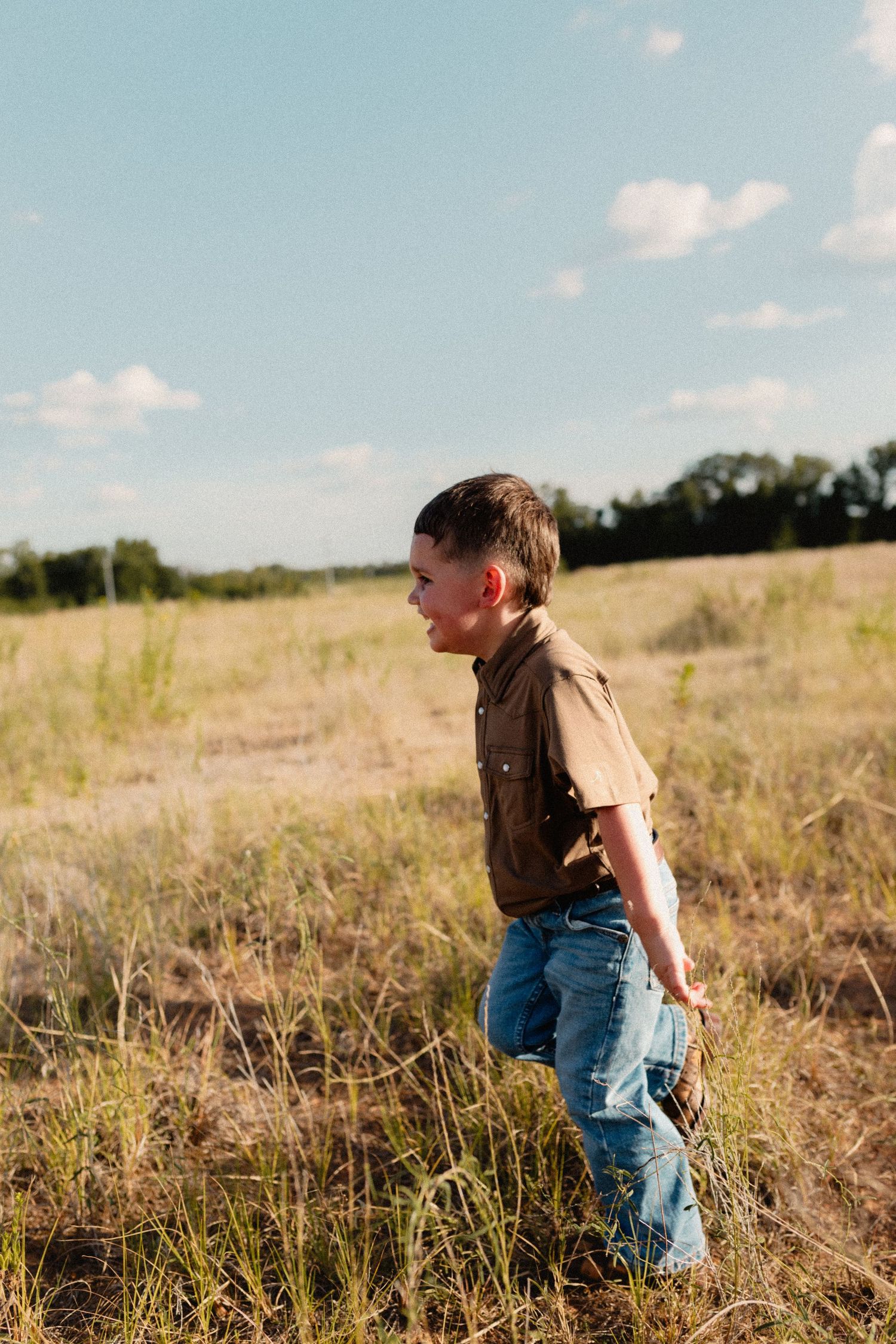 A young boy in a brown shirt and jeans walks through a grassy field under a blue sky.