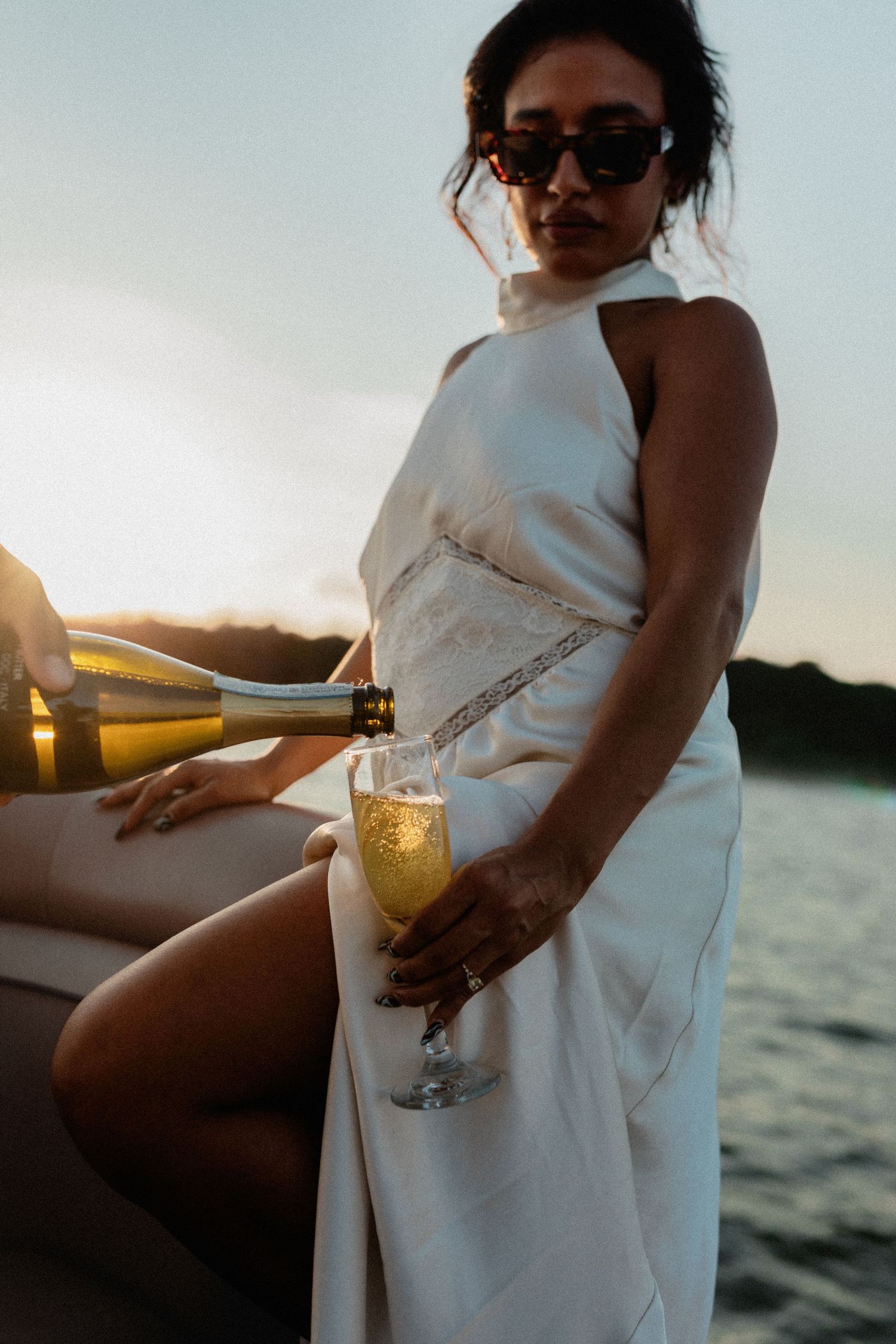 Woman in white dress and sunglasses receiving champagne pour on boat at golden hour.