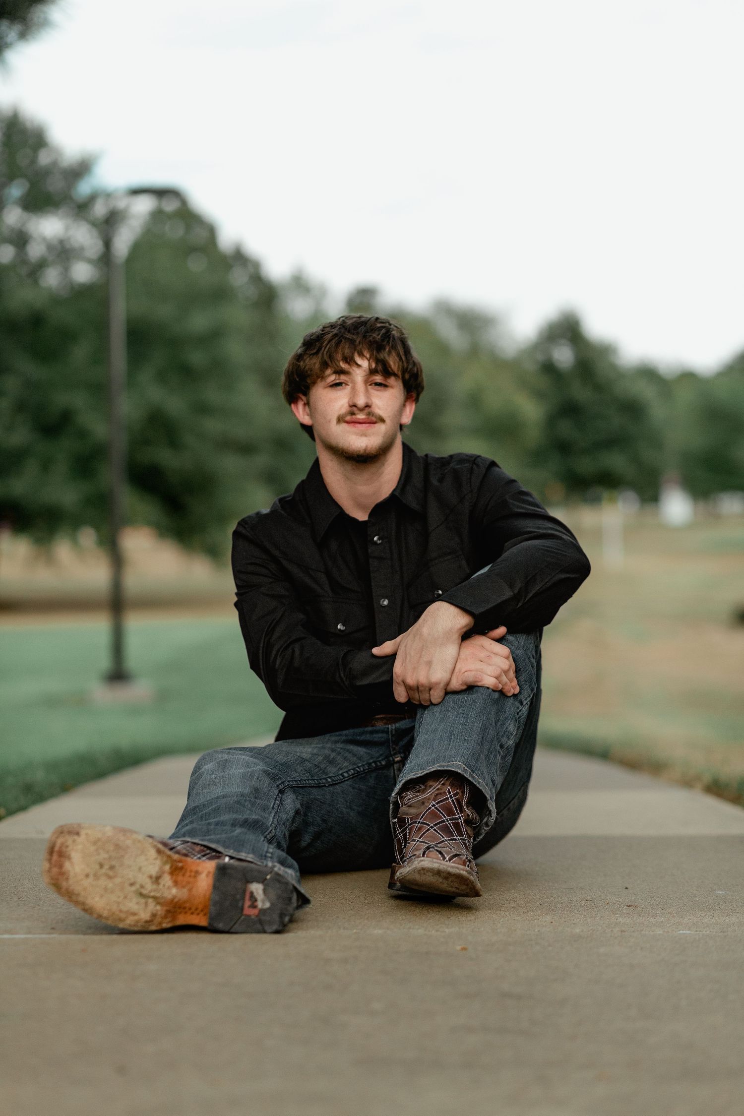 Smiling young man in black shirt sitting casually on pathway in outdoor park.