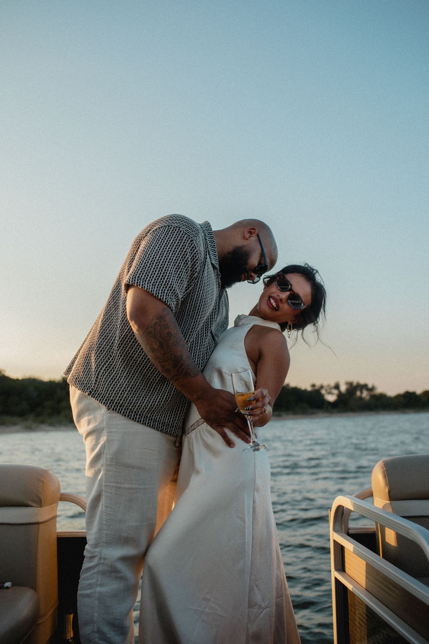 Tattooed man embracing laughing woman in white dress on boat deck at golden hour sunset.