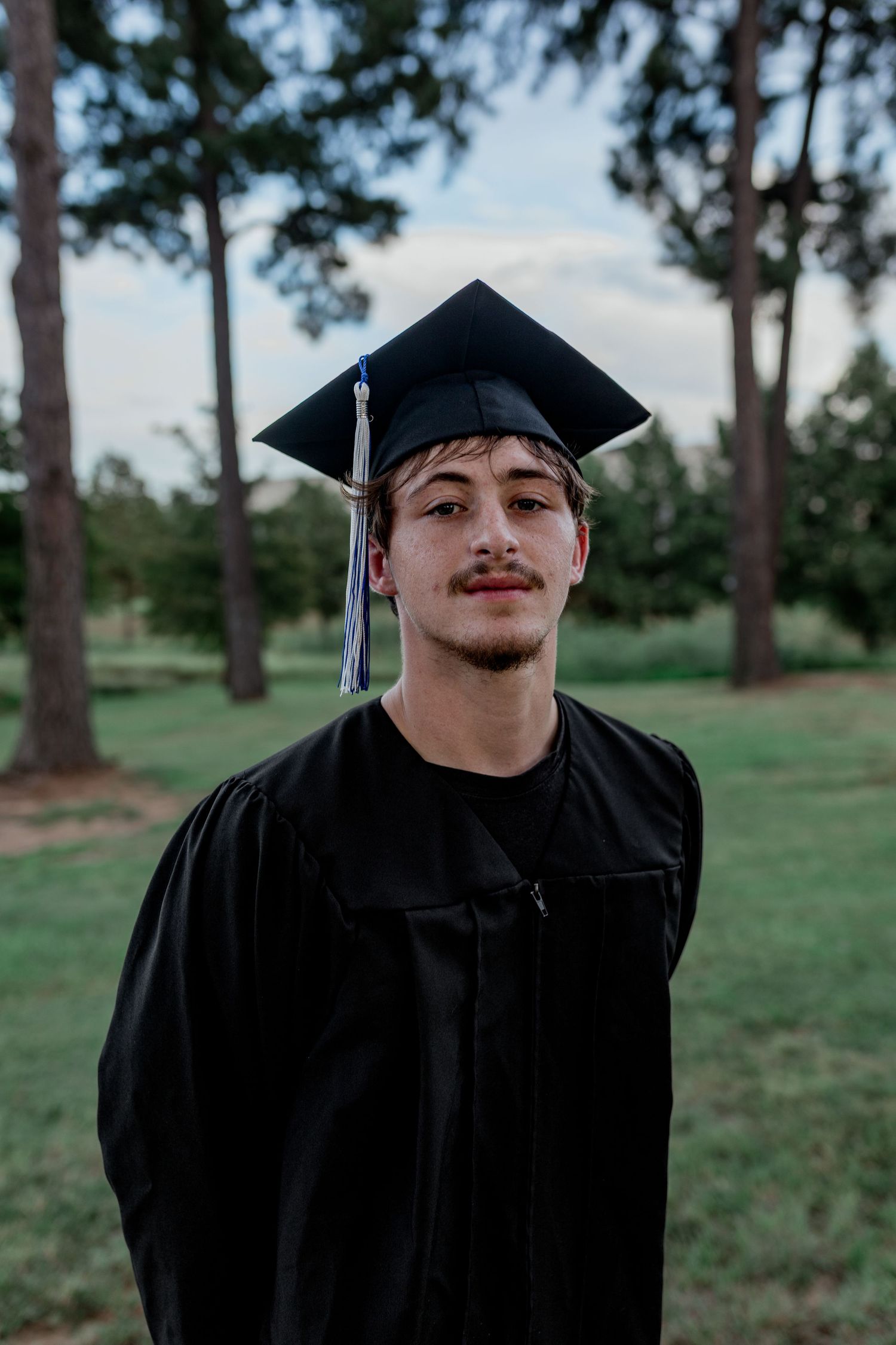 Young man in black graduation cap and gown poses confidently outdoors in a green park setting.
