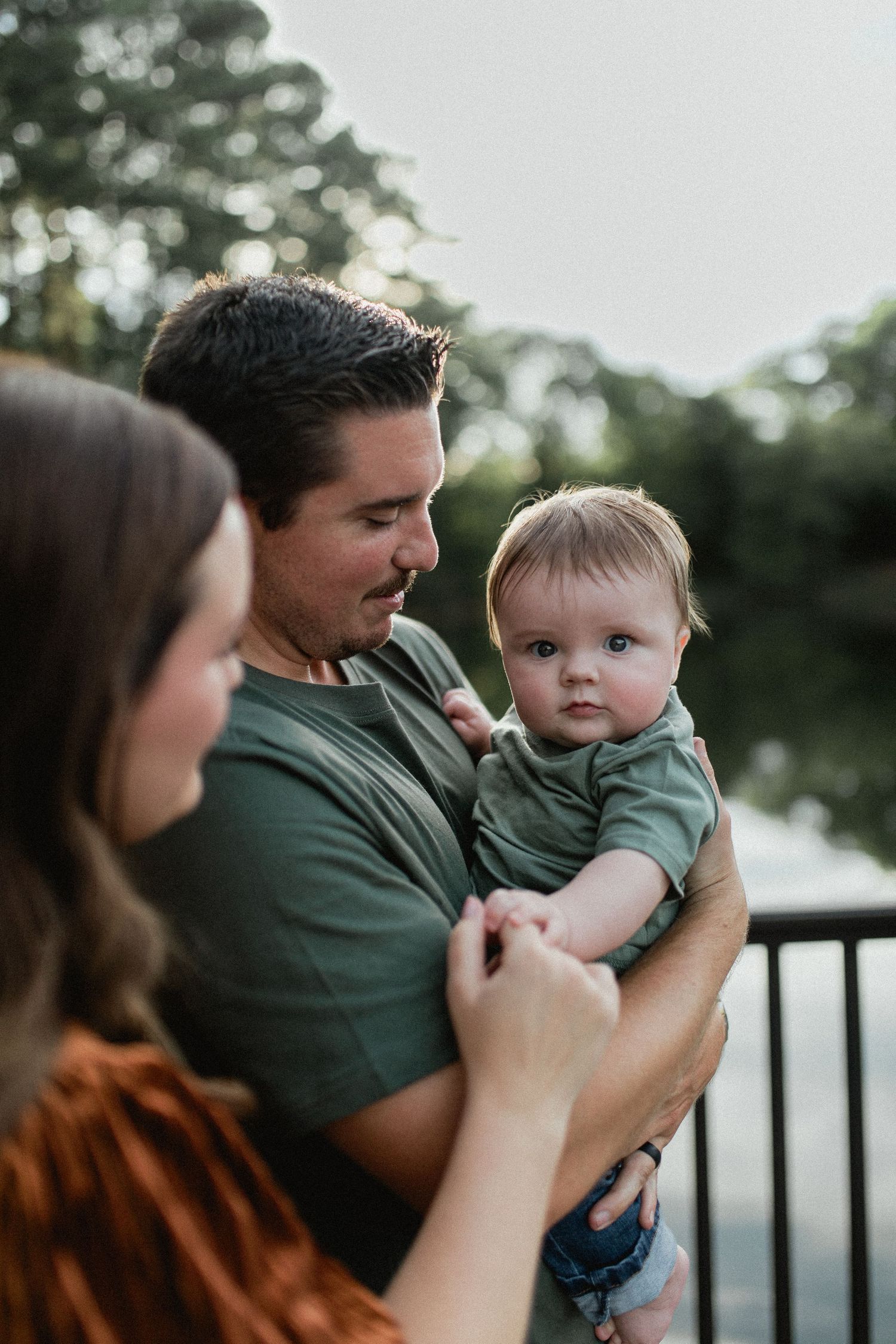 A father in a green shirt holds his baby boy while the mother reaches out near a lakeside railing.