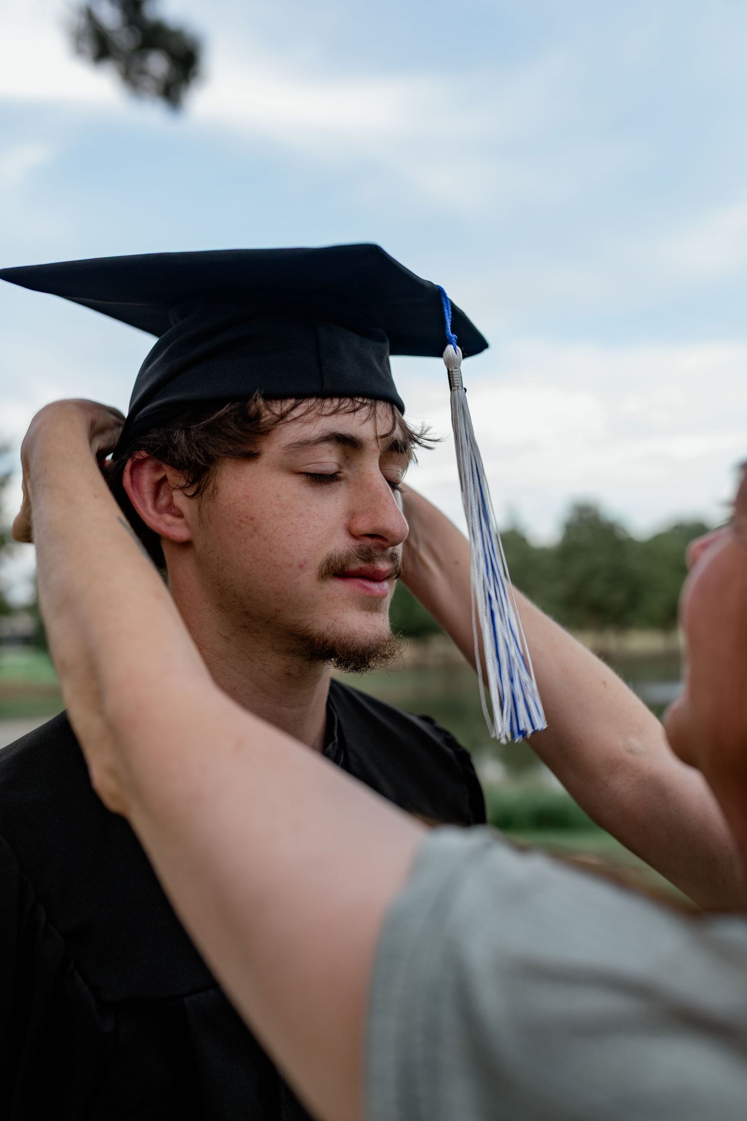 Woman placing graduation cap on young mans head outdoors under cloudy sky.