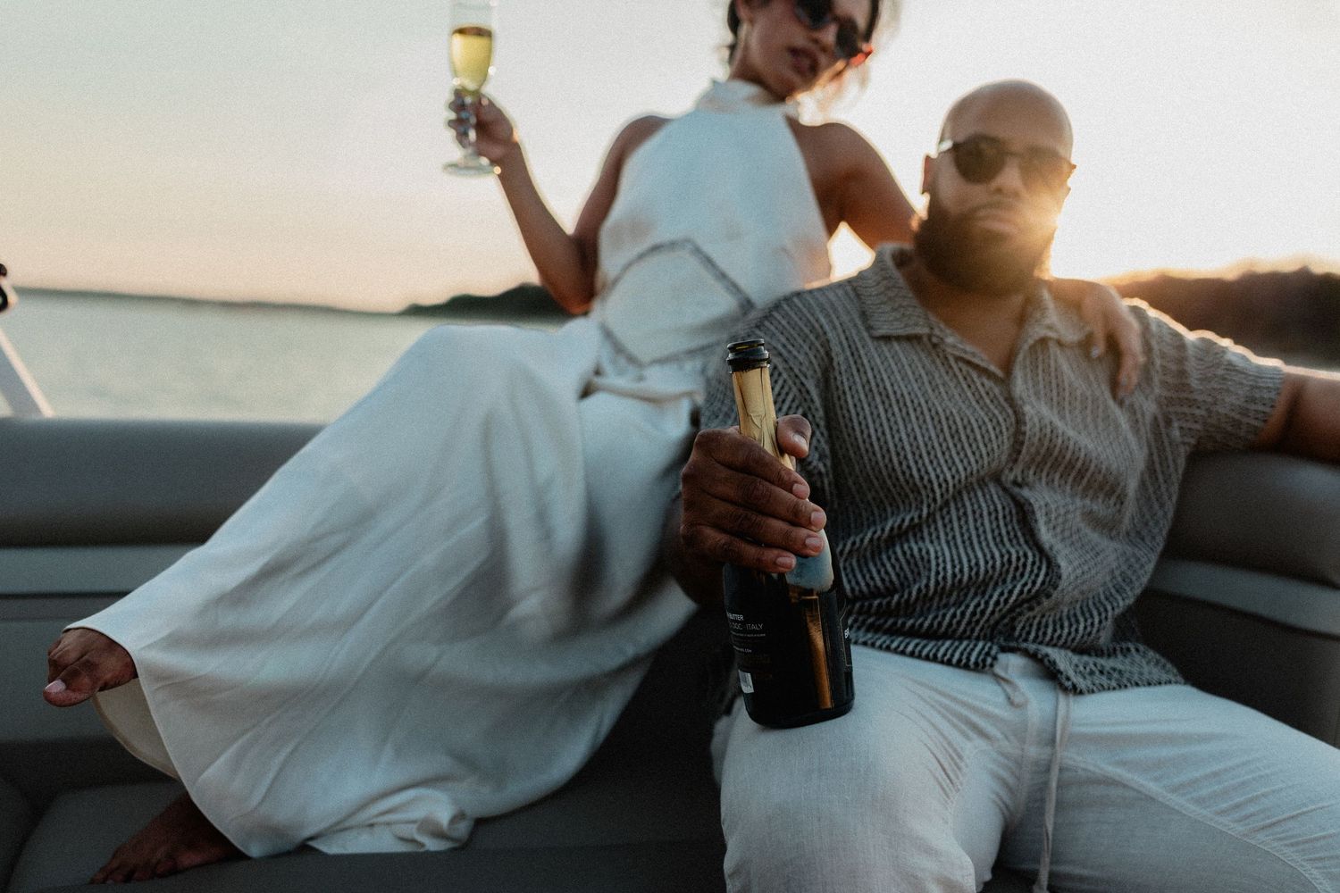 Couple on boat at sunset, man holding champagne bottle, woman in white dress standing.