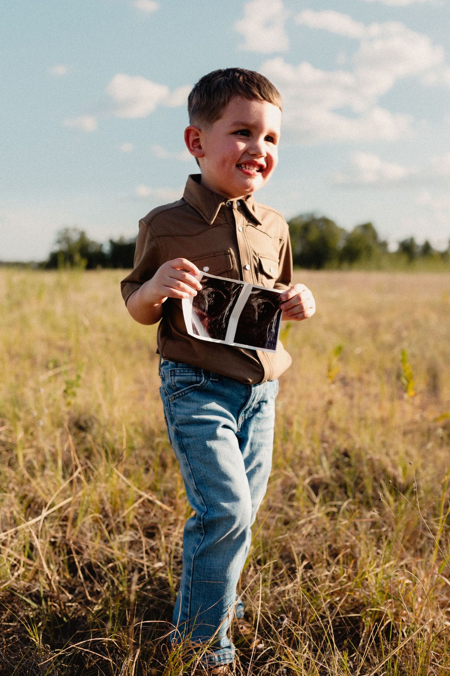 A happy young boy in jeans and brown shirt holds an ultrasound image in an open field.