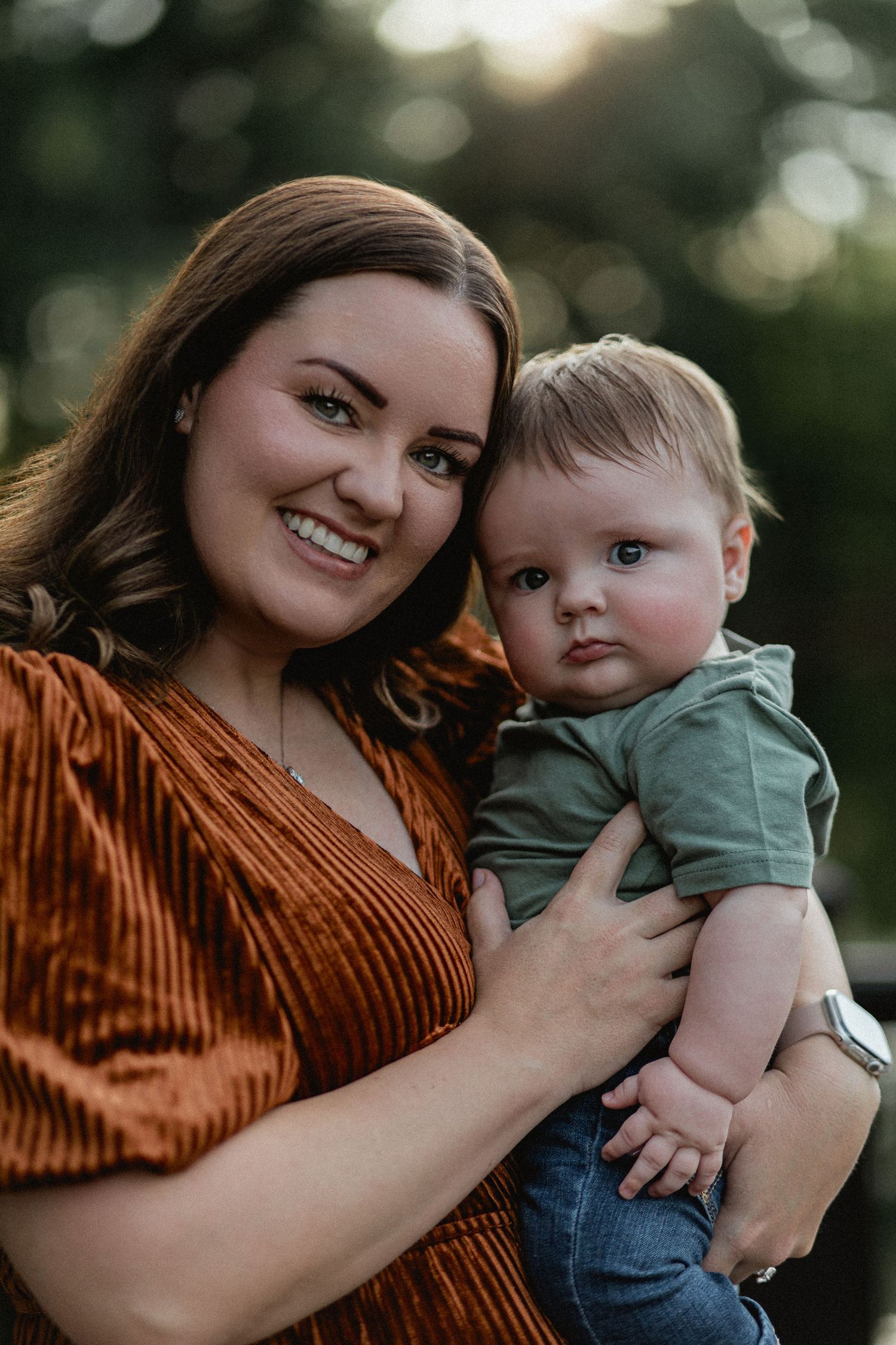 A smiling mother in a rust velvet dress holds her baby boy close in warm evening light.