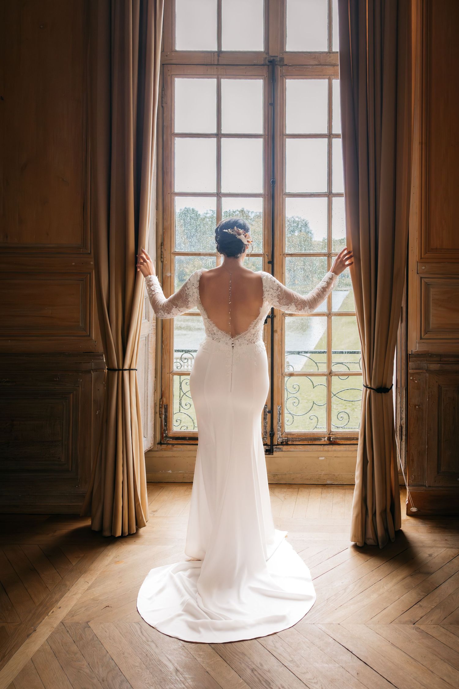La mariée en robe de mariée en dentelle sans dos élégante se tient devant une grande fenêtre de château avec des rideaux.