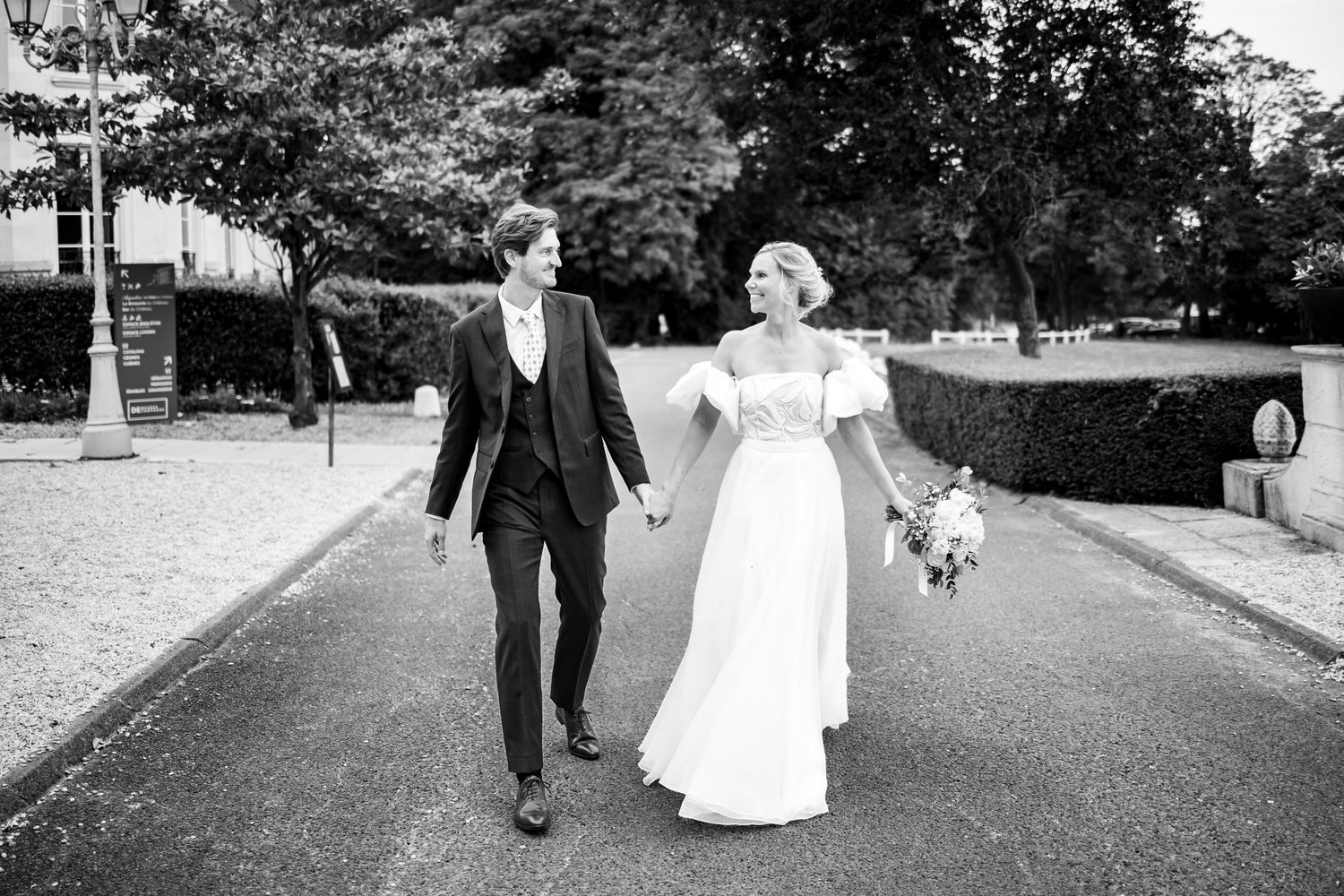 Photo en noir et blanc d'une mariée et d'un mariant marchant main dans la main le long d'un chemin bordé d'arbres.