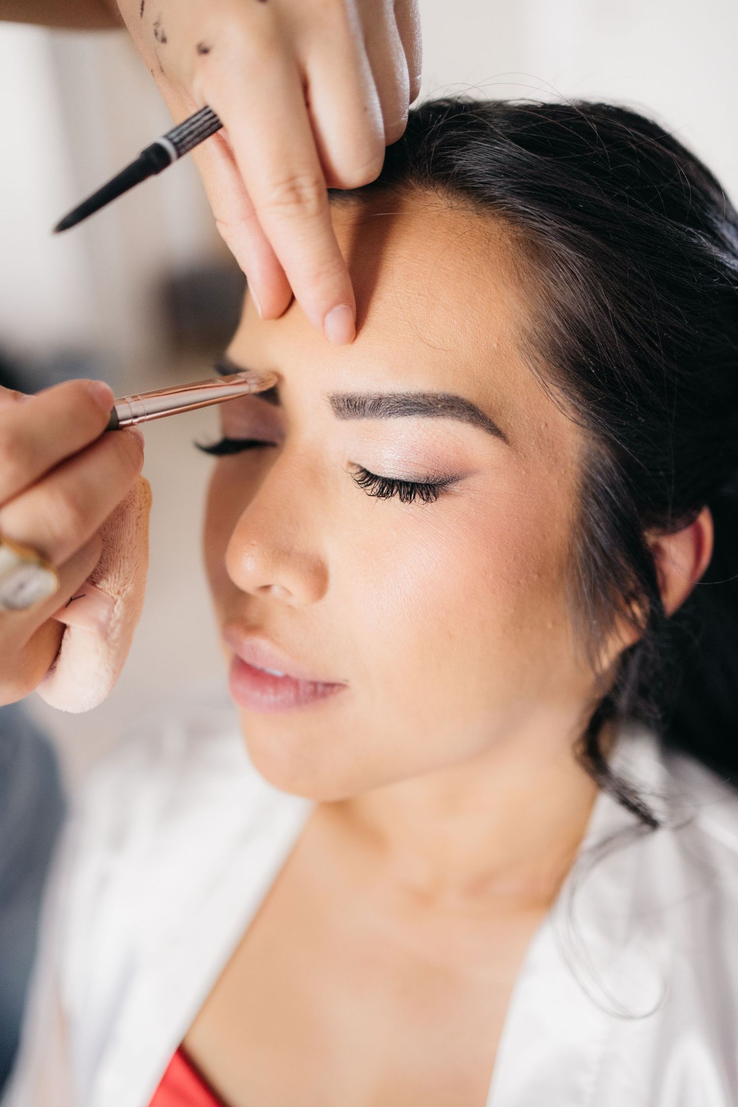 La maquilleuse applique soigneusement le maquillage des sourcils à une mariée aux cheveux foncés portant une robe blanche avant son mariage.