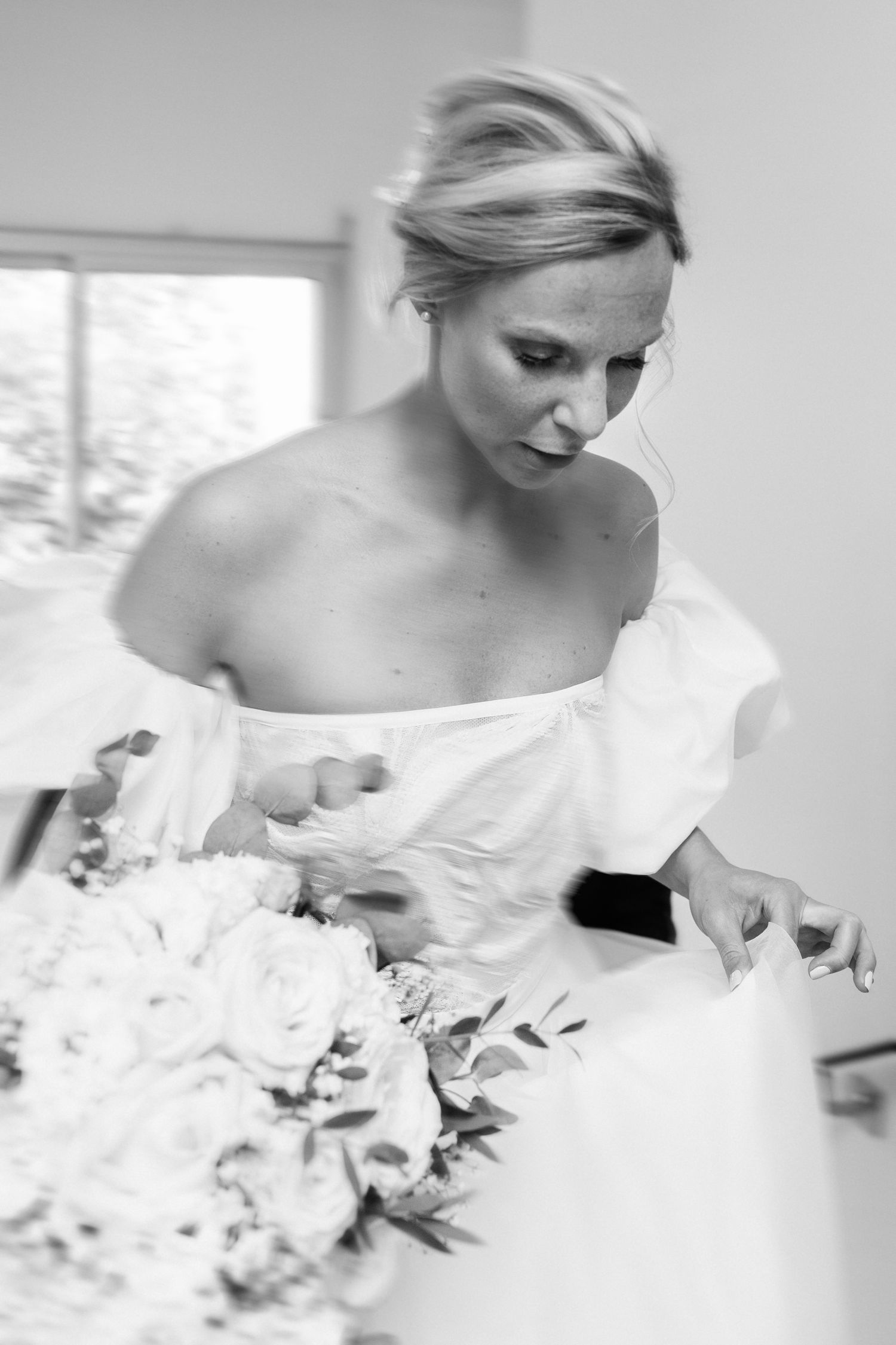 Photo en noir et blanc d'une mariée dans une robe à épaules ouvertes disposant de son bouquet de mariée de roses blanches.