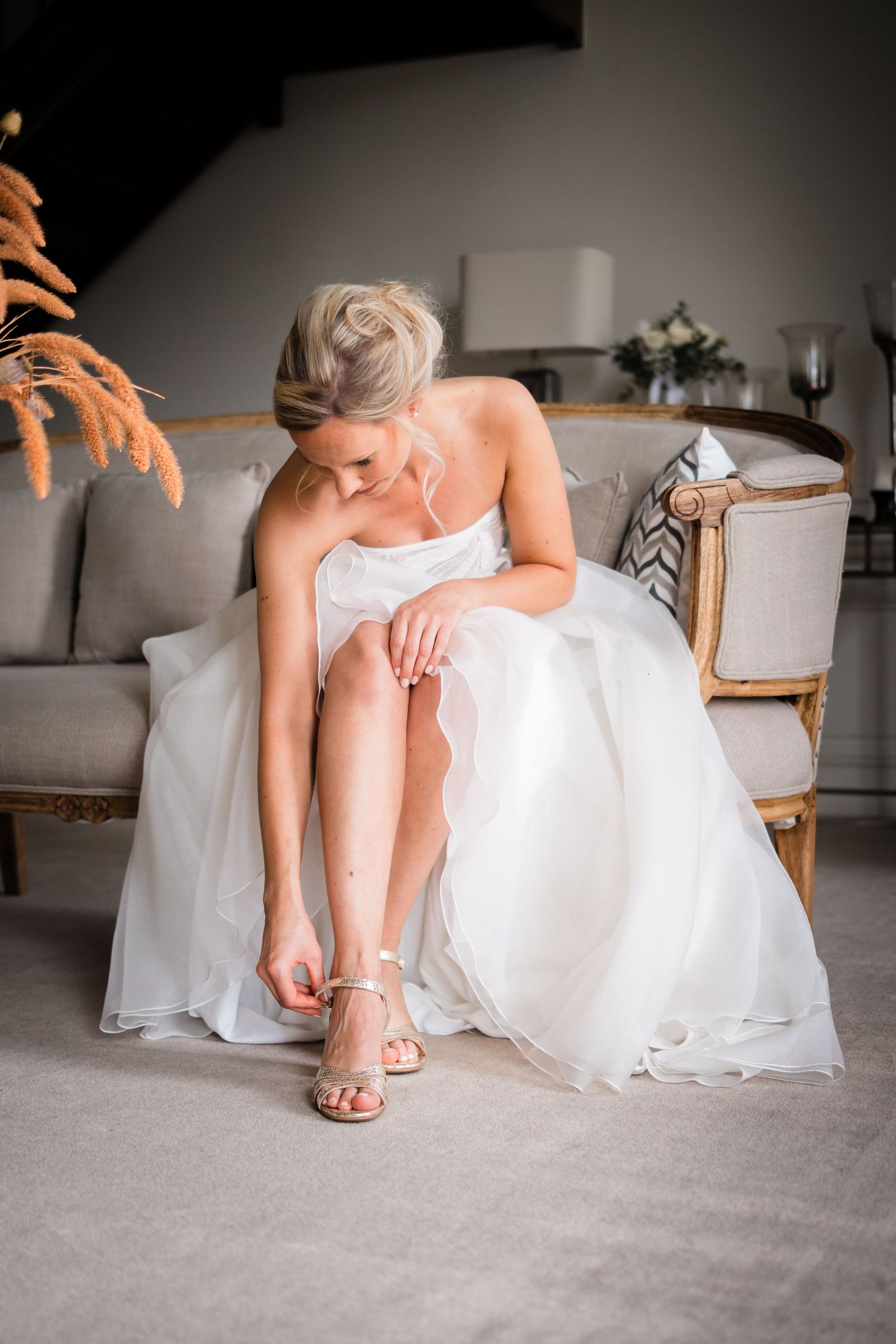 La mariée blonde en robe de bal blanche s'assoit sur un canapé élégant en attachant ses talons de mariage.