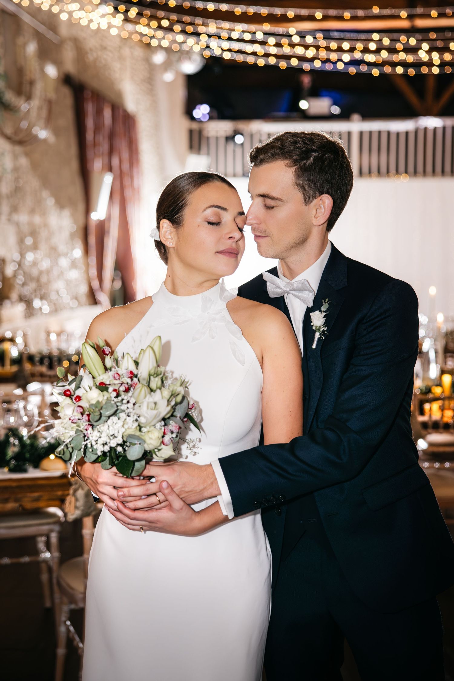 Bride and groom embrace tenderly at wedding reception, bride holding white floral bouquet, groom in navy suit with boutonniere.