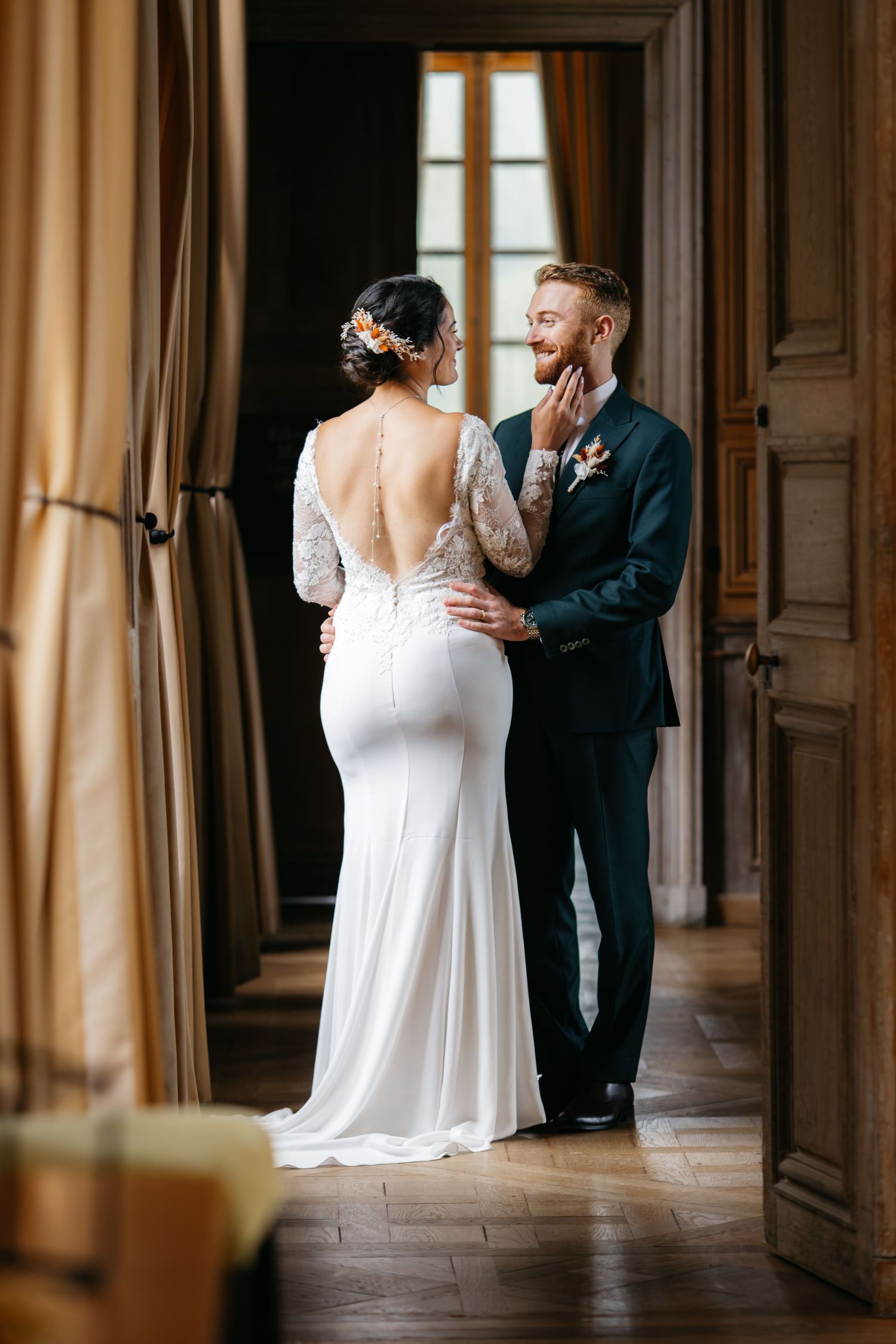 La mariée dans une robe de mariée en dentelle sans dos touche tendrement le visage du marié dans une porte de château vintage.