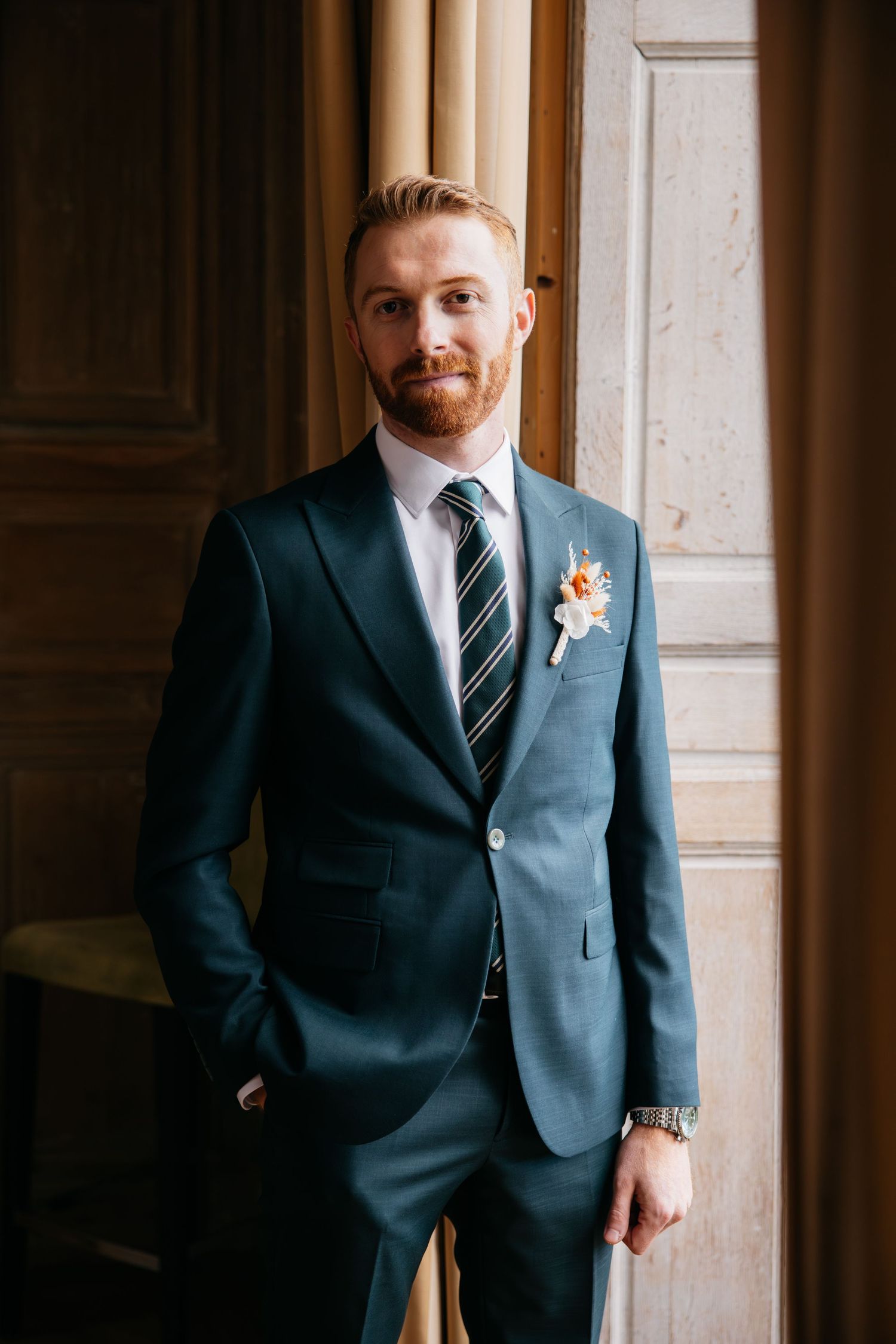 Beau mari roux barbu en costume vert émeraude avec boutonnière posant avec assurance devant une porte en bois.