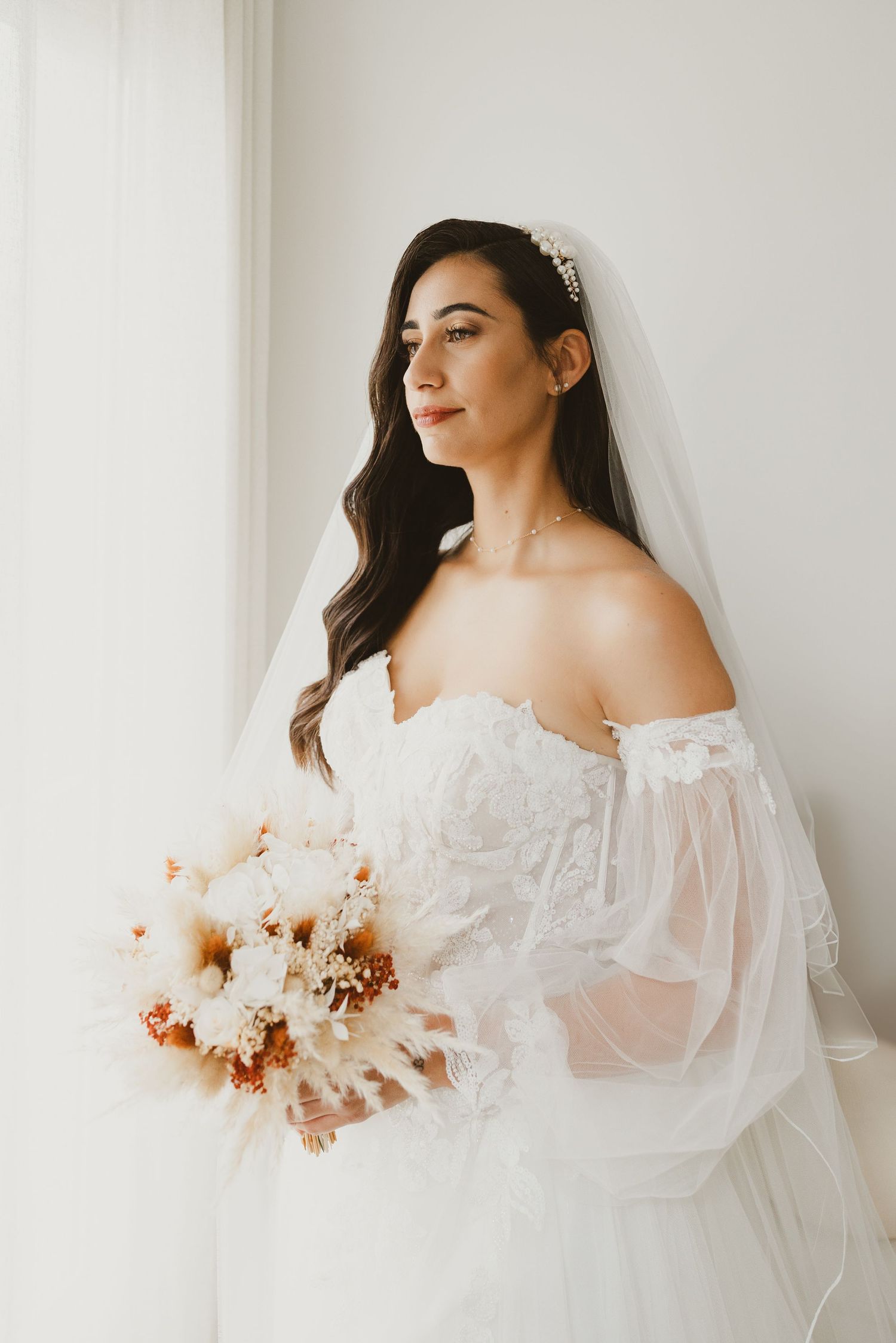Élégante mariée en robe de mariée en dentelle à épaules ouvertes tenant un bouquet bohème avec des fleurs séchées par la fenêtre.