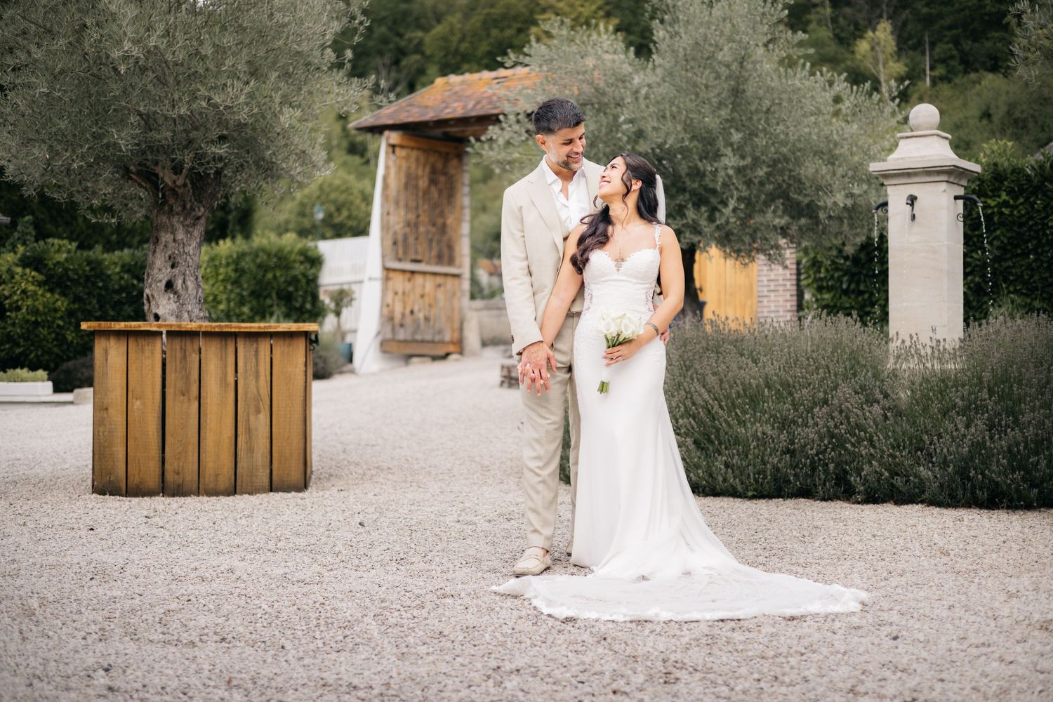 La mariée en robe blanche ajustée et le marié en costume marron se tiennent ensemble dans un jardin ensoleillé avec des oliviers.