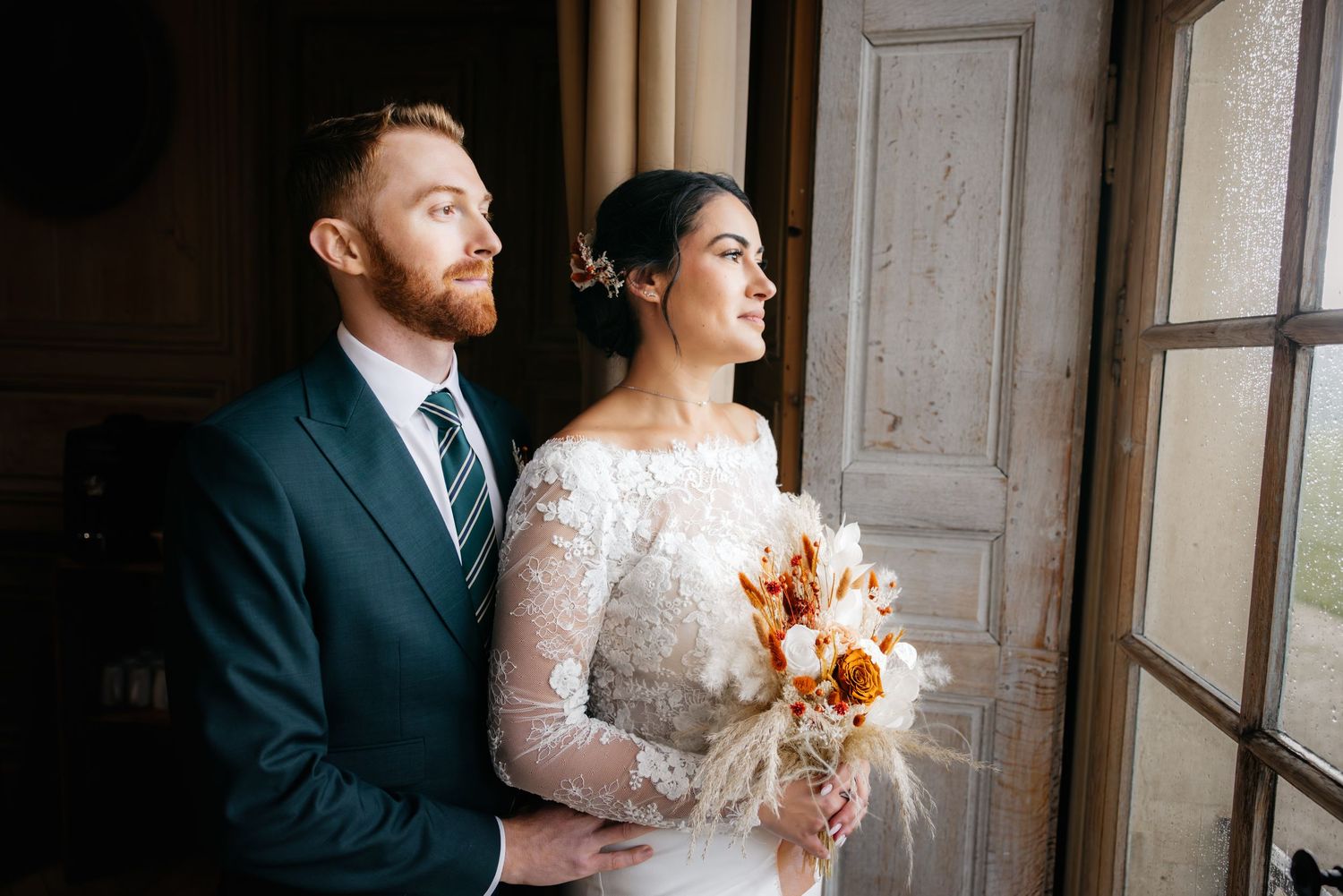 La mariée et le marié regardent par la fenêtre ensemble, la mariée tient un bouquet bohème dans une robe de mariée en dentelle.