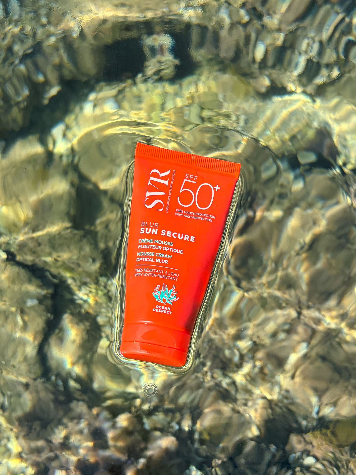 Tube de crème solaire SPF 50+ posé sur des galets dans une eau translucide — photographie de produit en extérieur