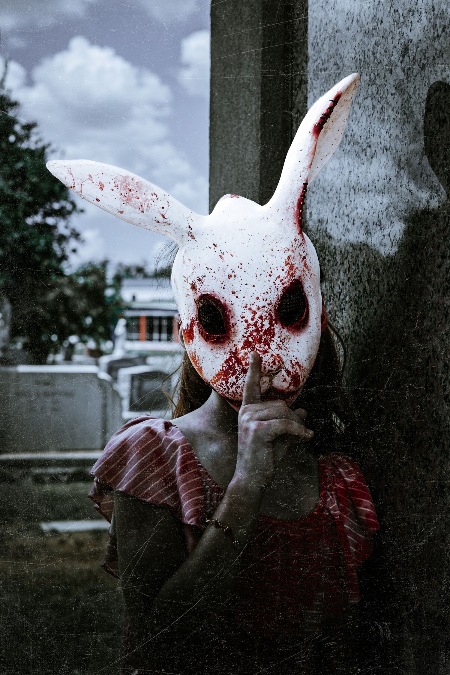 Person in a blood-splattered white rabbit mask holds a finger to the lips beside a gravestone in a cemetery under a cloudy sky.