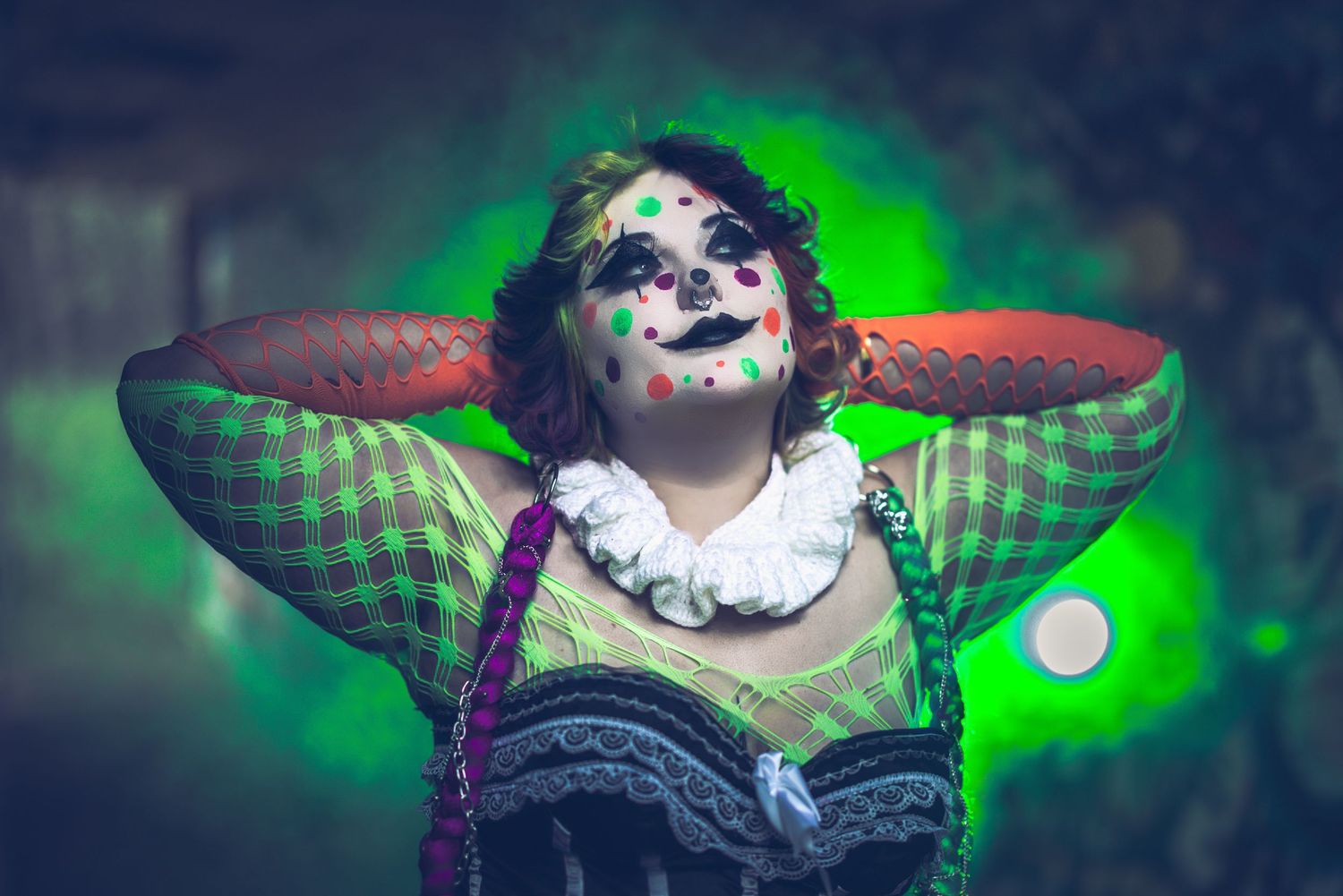 Woman in colorful clown makeup and white ruffled collar poses with arms raised behind her head against a glowing green background.