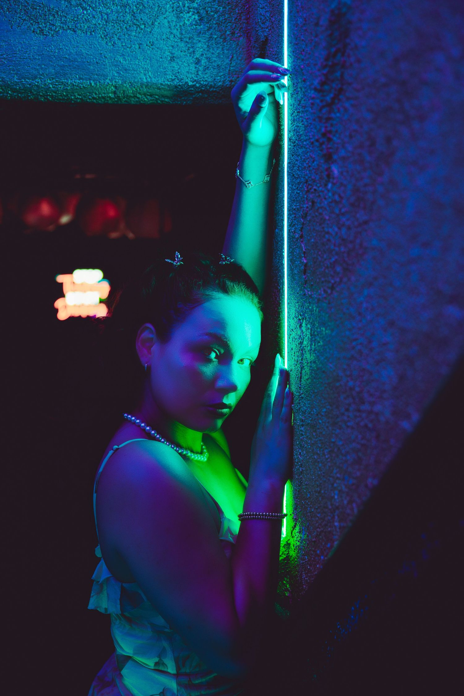 Young woman leaning against a wall illuminated by green and blue neon lights at night.