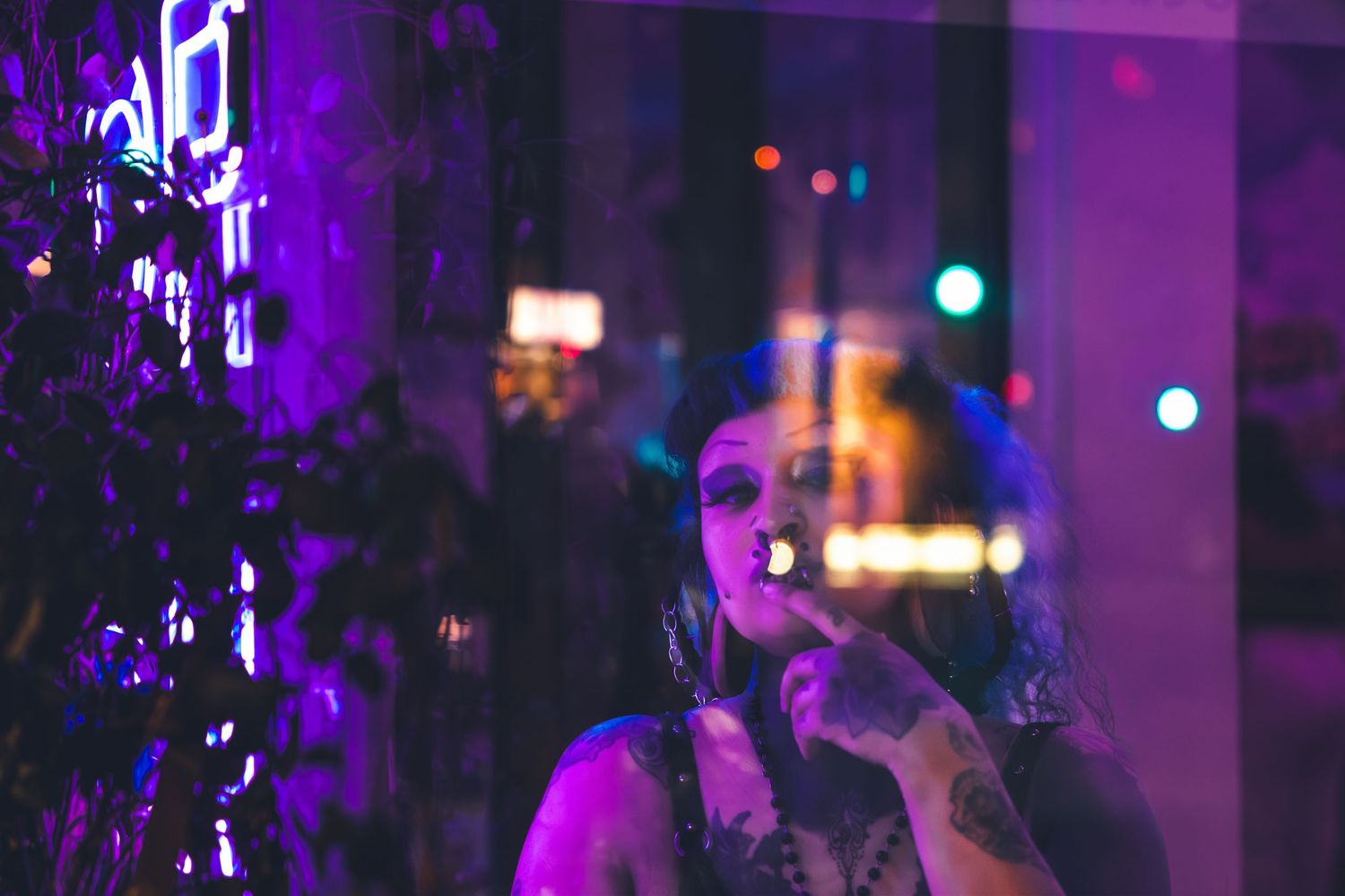 Tattooed woman smoking cigarette bathed in purple neon light with city reflections at night.