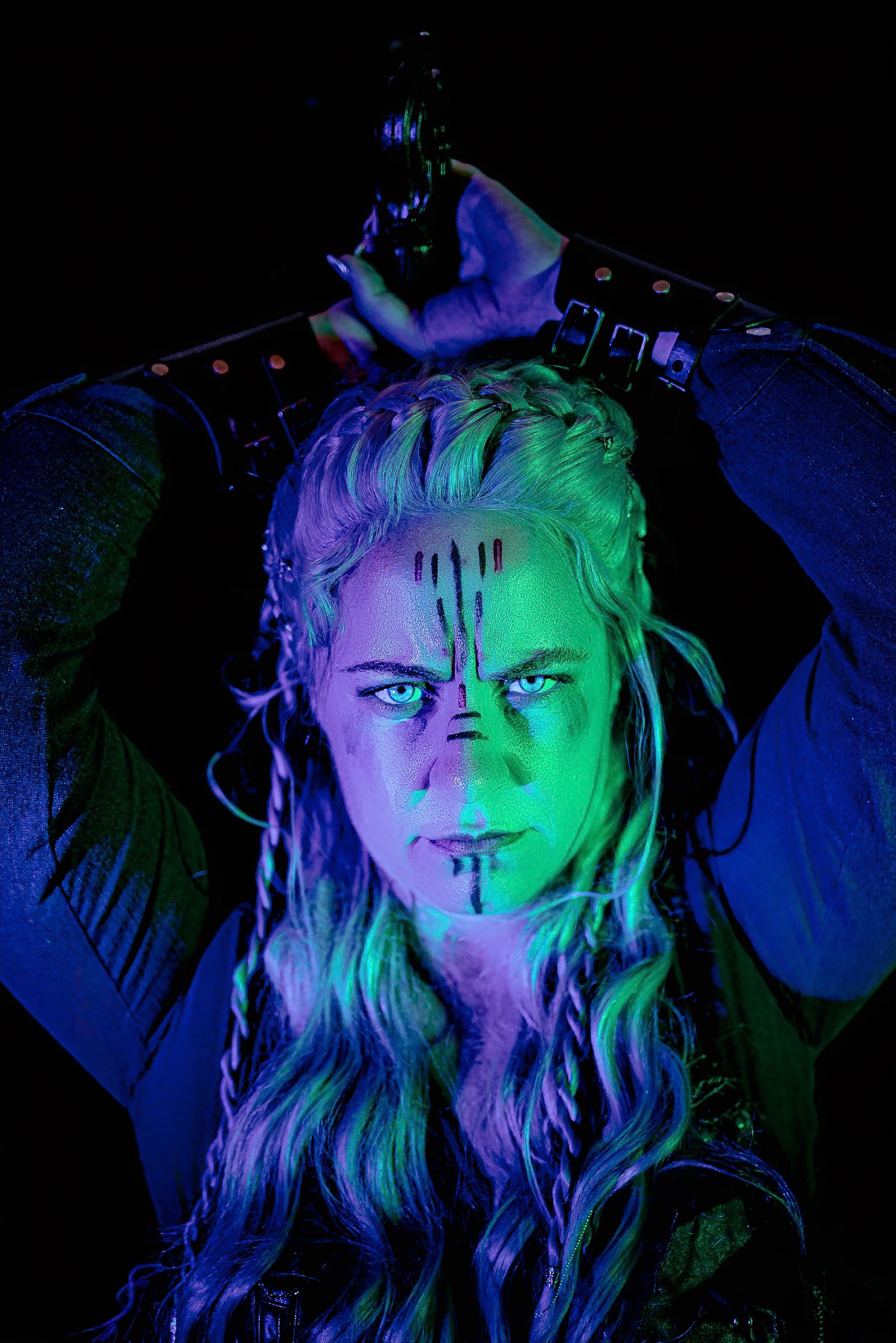 Warrior-style portrait of a braided blonde woman with dark face paint, hands raised overhead, lit by dramatic blue and green light.