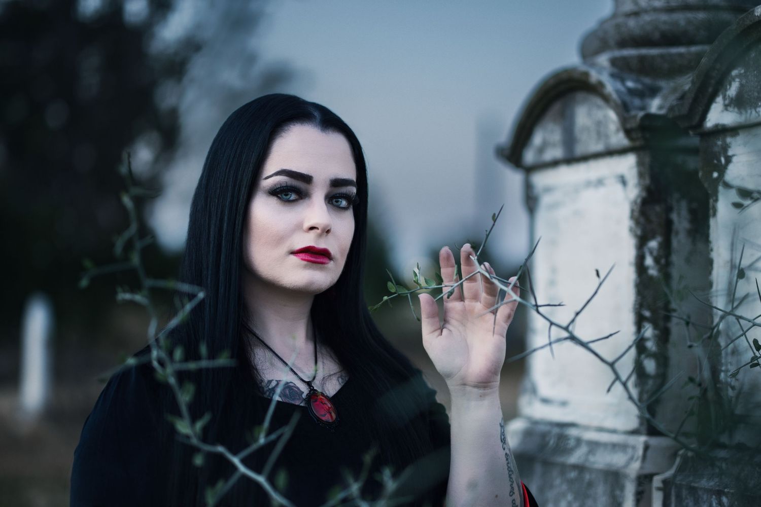 Gothic woman with black hair and red lips poses near a stone grave in a dark cemetery setting.