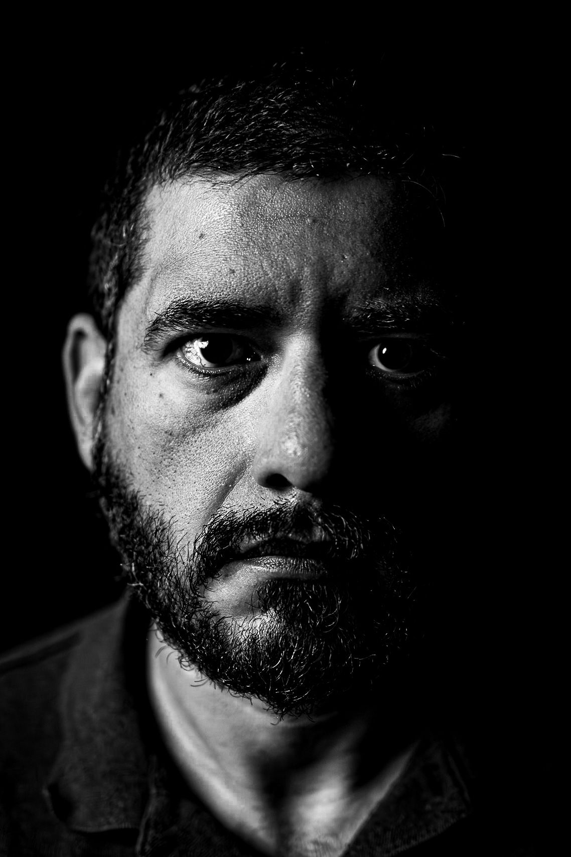 Dramatic black and white close-up portrait of a bearded man with intense eyes against a dark background.