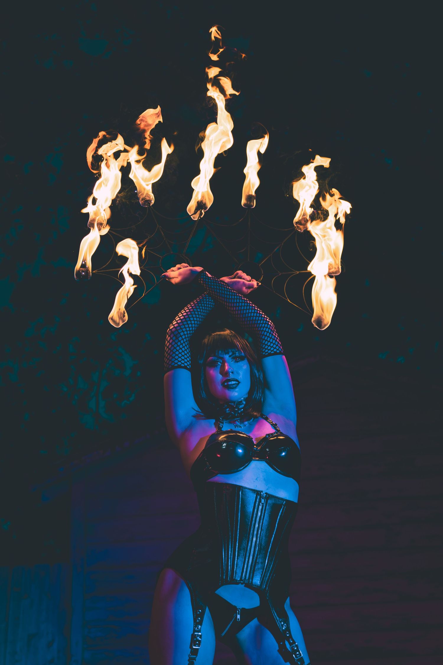 Fire performer in a black corset and fishnet sleeves raises her arms overhead while flaming fans burn against a dark night background.