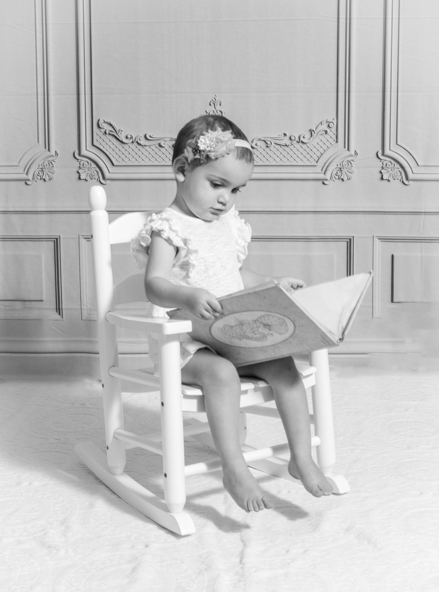 Black and white portrait of a toddler sitting in a white rocking chair reading a book by Tiny Toes to Tassels Photography in Springtown, TX