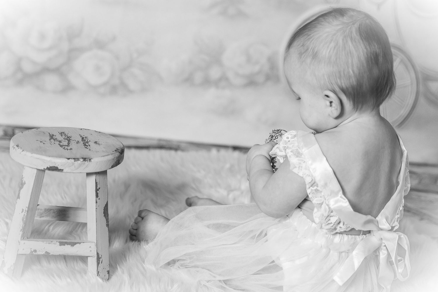 Baby girl sitting on a small wooden stool wearing a white dress in a studio portrait session by Tiny Toes to Tassels Photography
