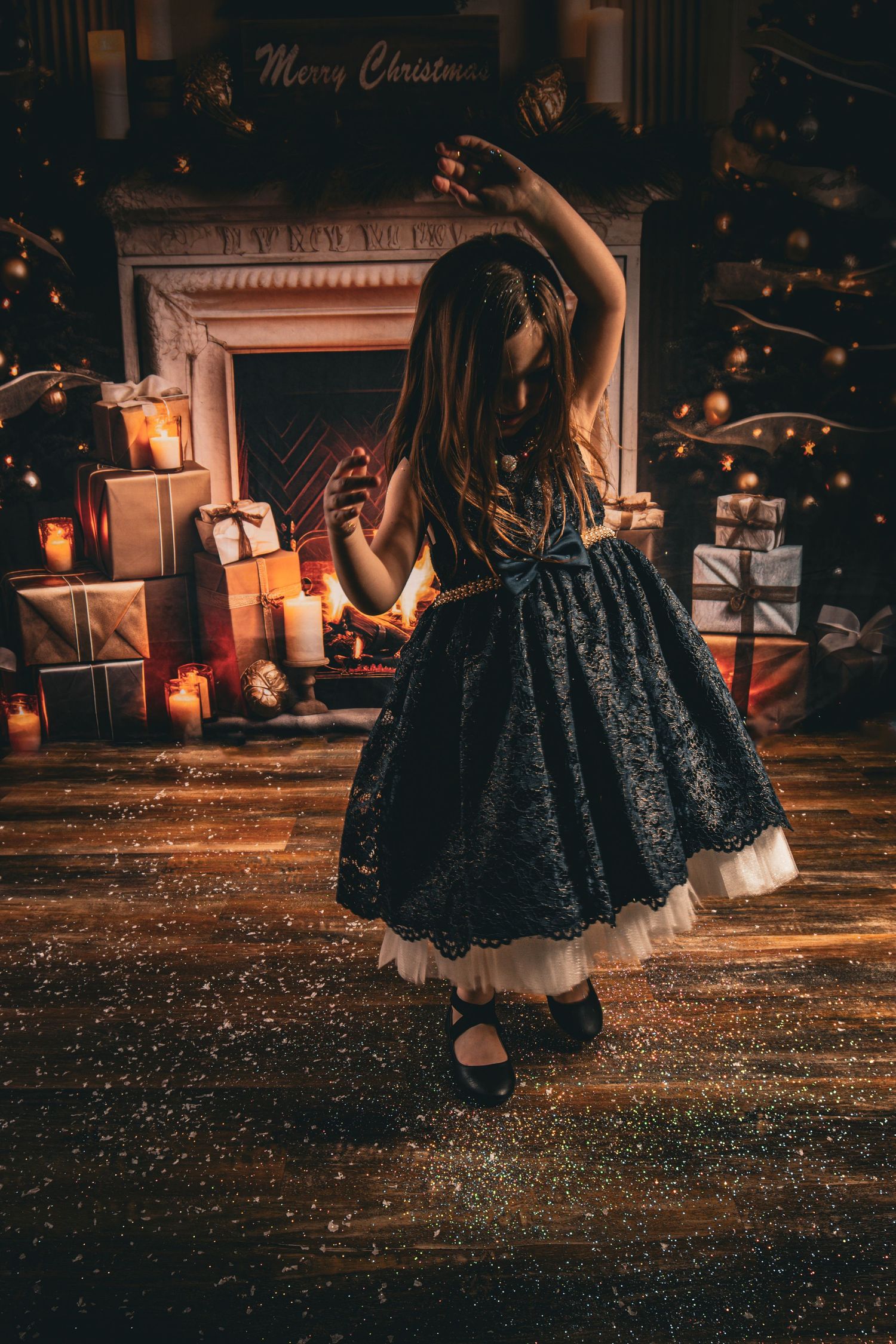 Young girl in a dark holiday dress standing in front of a Christmas fireplace and tree backdrop by Tiny Toes to Tassels Photography