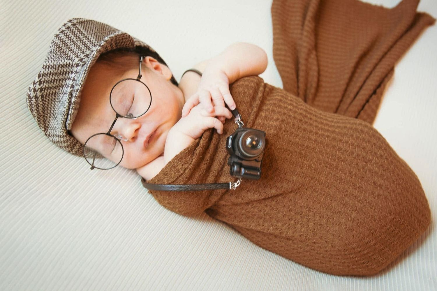 Newborn baby wearing adorable round glasses and a newsboy cap wrapped in brown knit by Tiny Toes to Tassels Photography