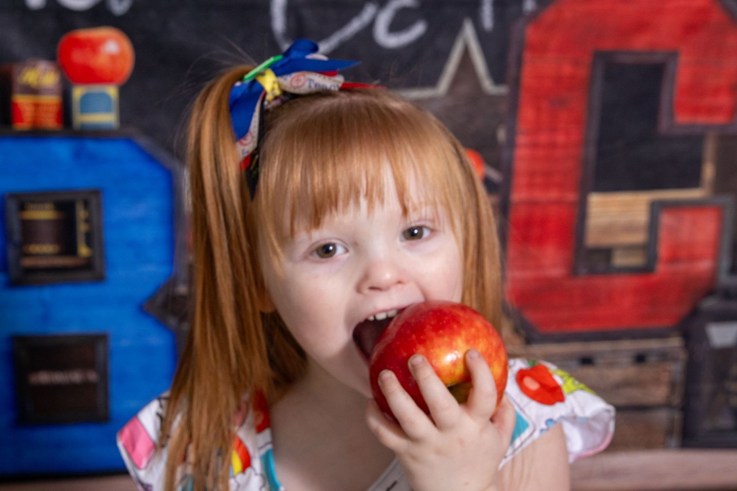 Toddler girl eating a red apple in front of a school-themed backdrop by Tiny Toes to Tassels Photography in Springtown, TX