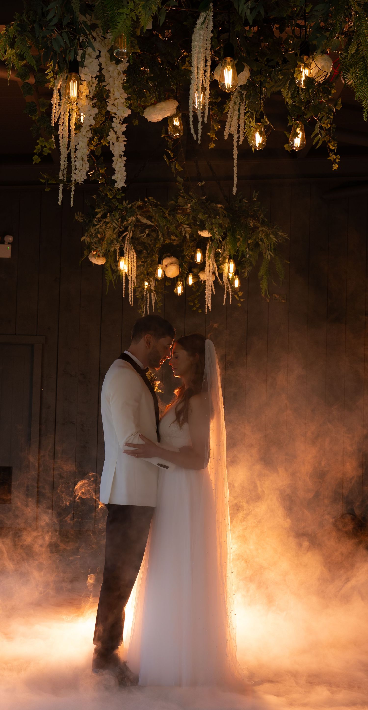 Bride and groom sharing an intimate embrace beneath warm lights and atmospheric smoke at their wedding venue.