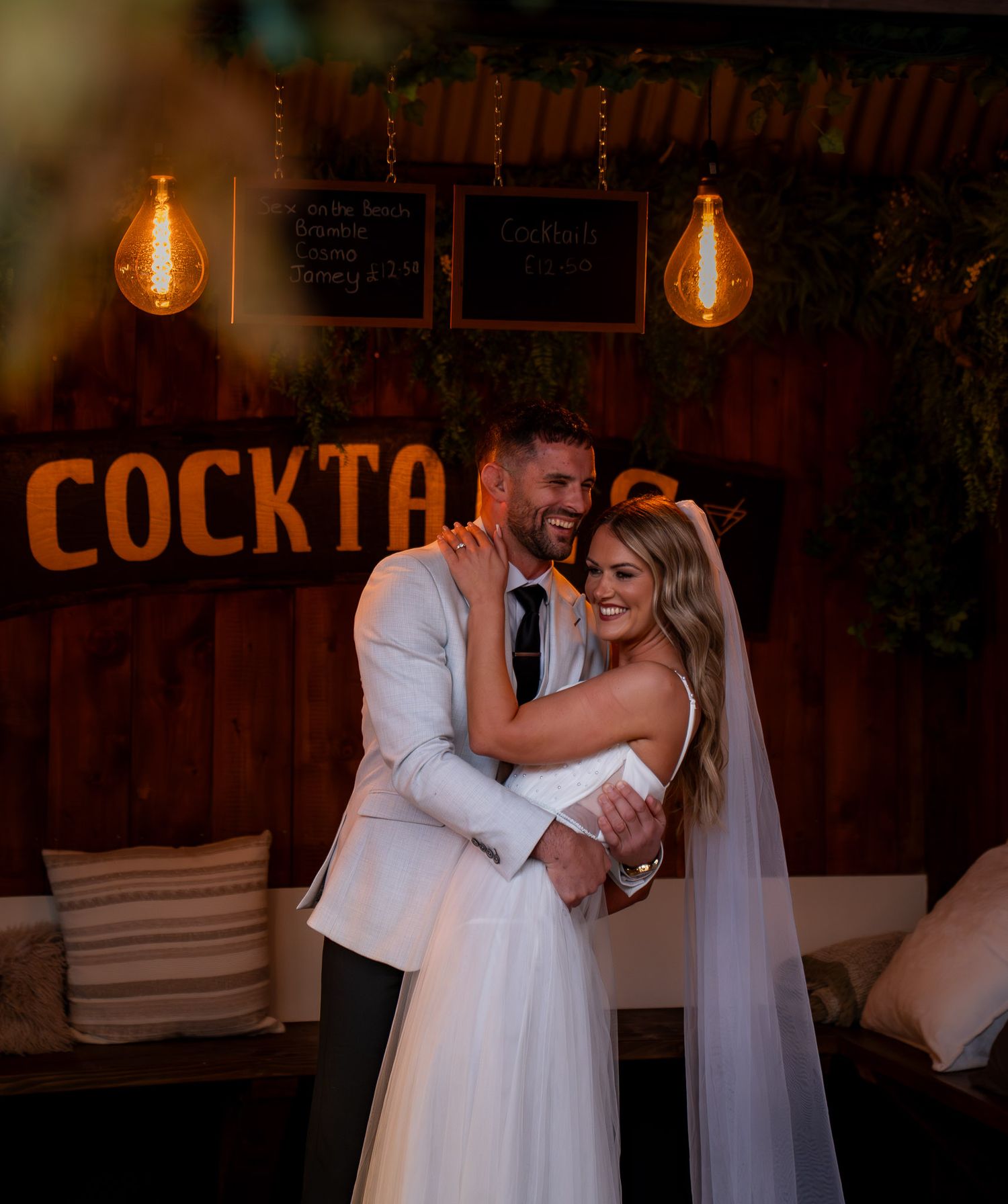 Bride and groom embracing beneath warm hanging lights in the wedding venue’s cocktail bar.