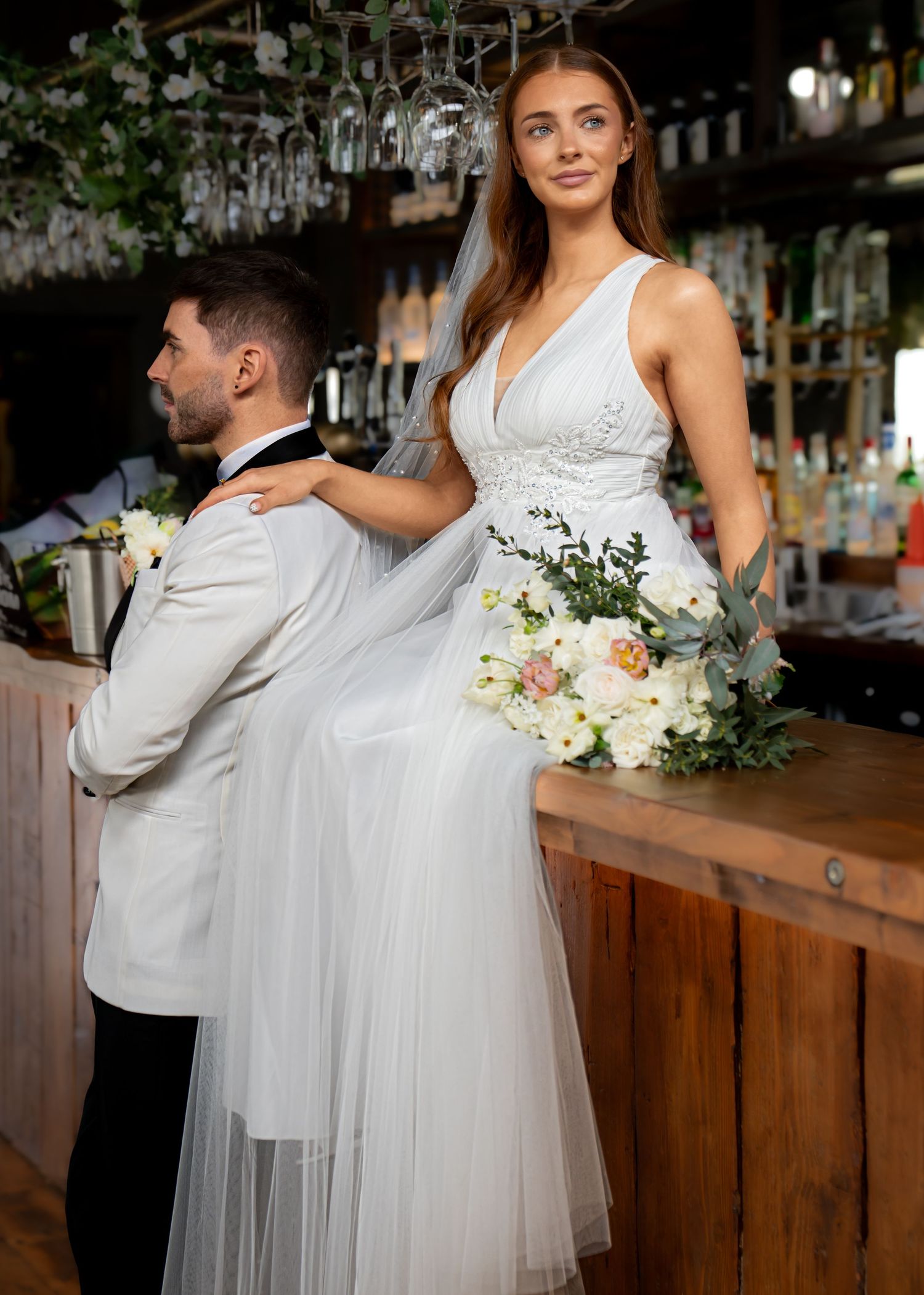 Bride and groom posing together at the wedding venue bar with her colourful bouquet beside them.