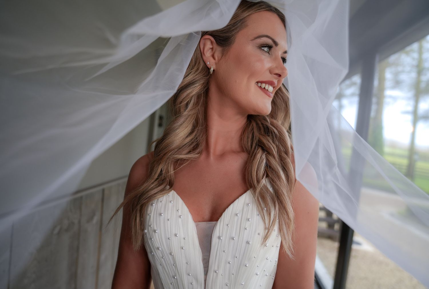 Smiling bride beside a window with her flowing veil framing her elegant beaded wedding dress.