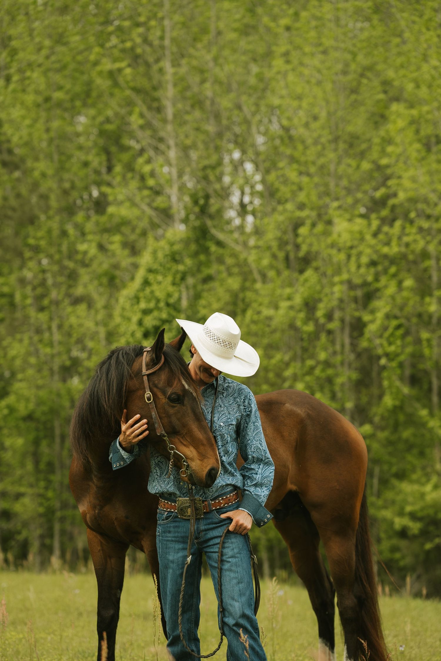 Equine portrait photography featuring a man looking at his horse in a green pasture surrounded by trees.