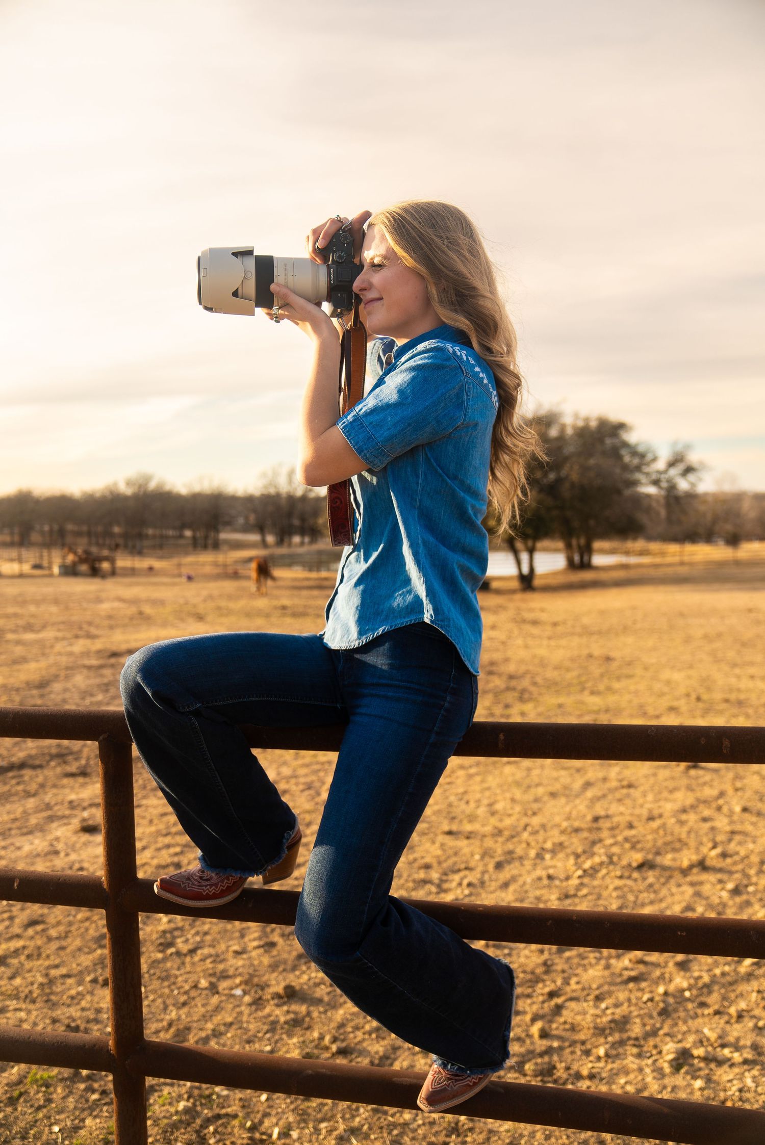 Behind-the-scenes portrait of photographer Joy Nabors sitting on a fence with her camera during a photo session.