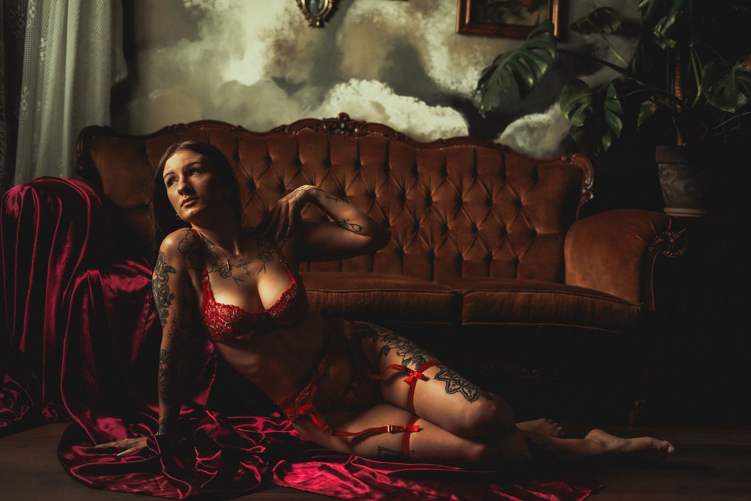 Tattooed woman in red lingerie posing elegantly beside vintage velvet sofa in dramatic lighting.