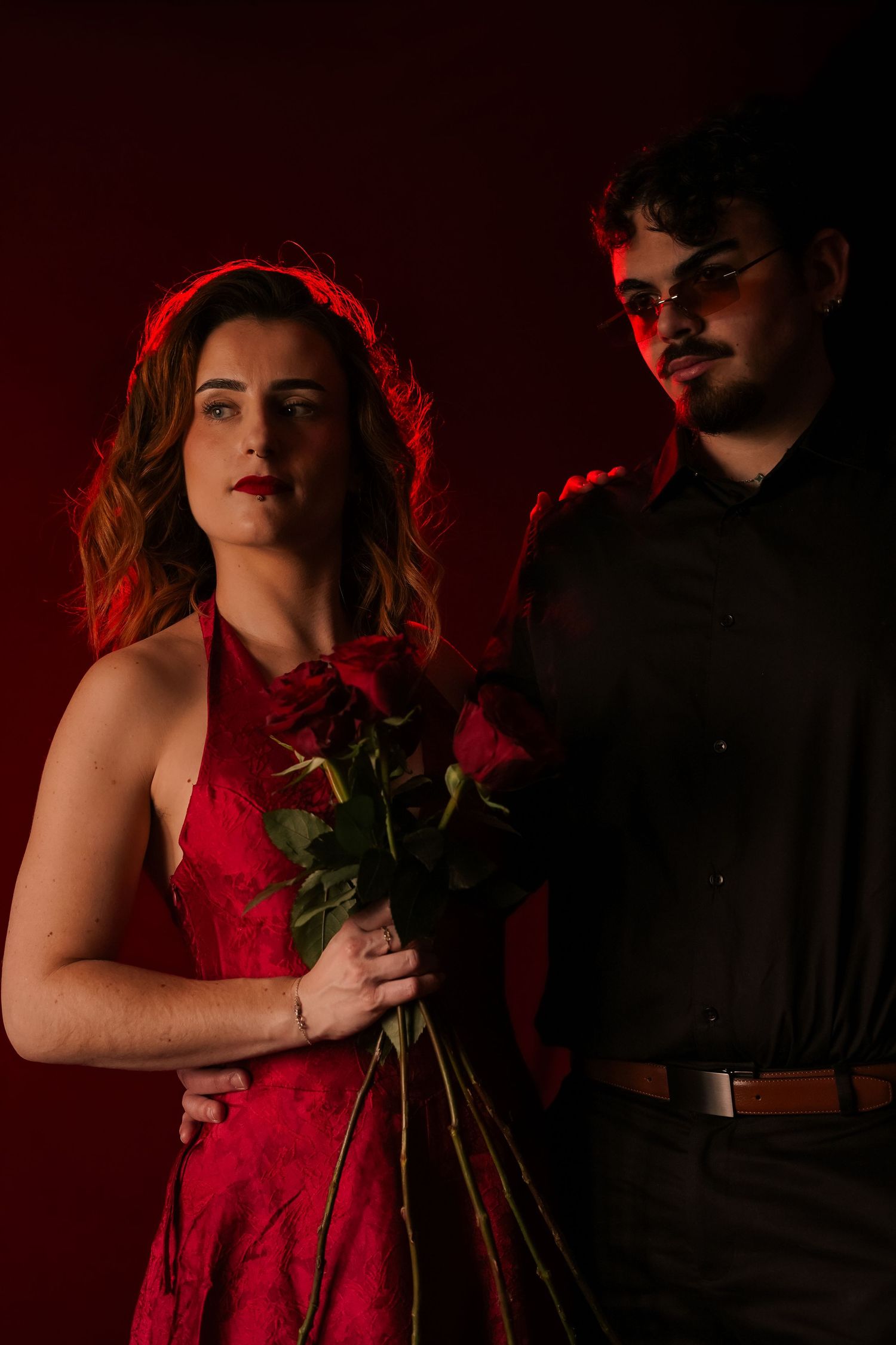 Woman in red dress holds roses while man in black shirt stands behind her in moody red light.