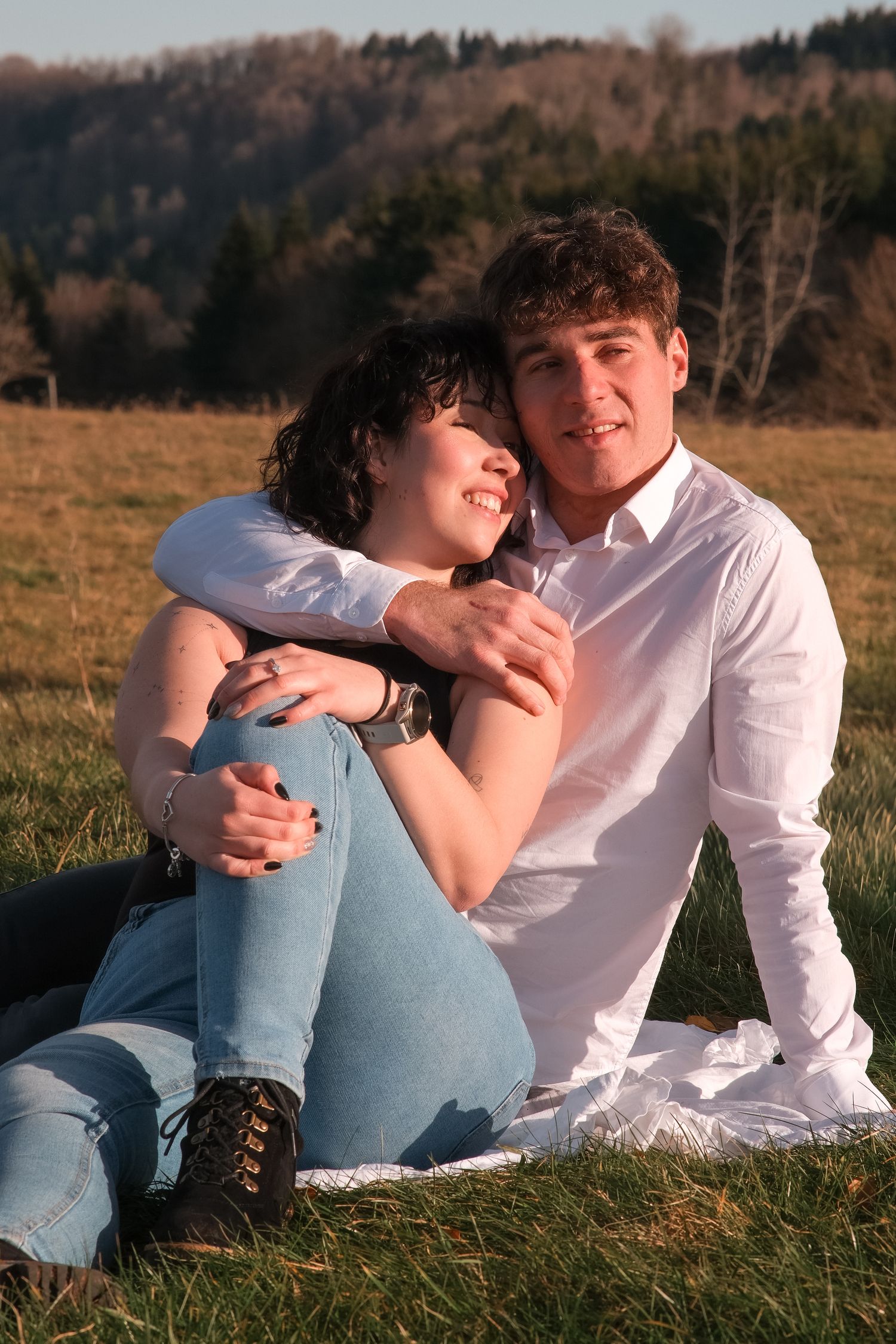 Young couple sitting on grass embracing warmly at golden hour in scenic meadow.