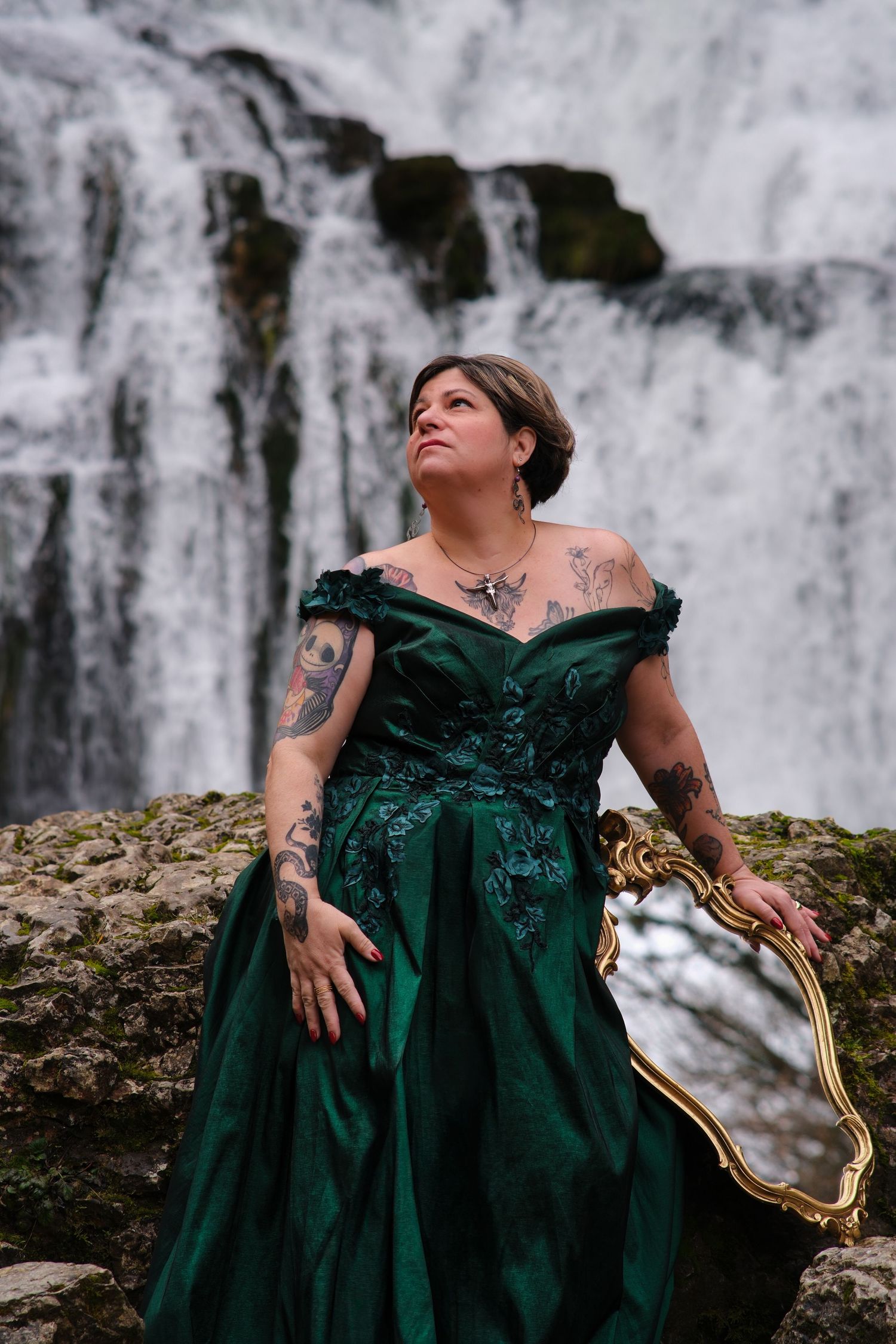 Tattooed woman in emerald green off-shoulder ballgown holds gold mirror before misty cascading waterfall.