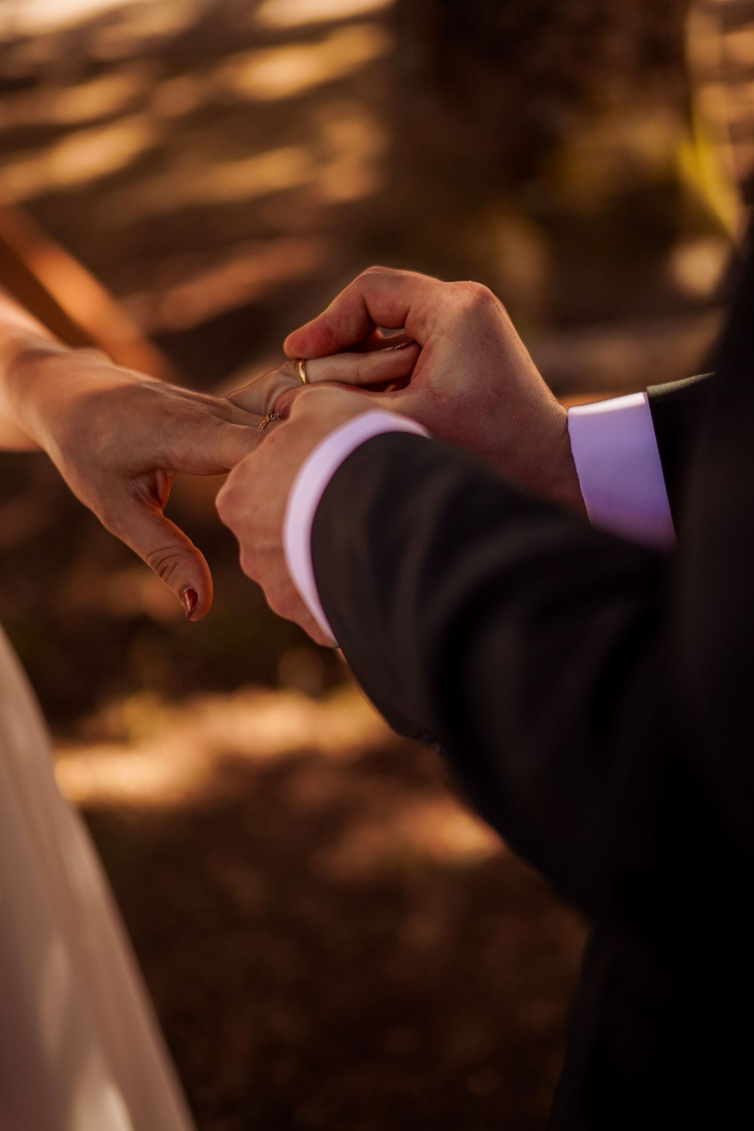 Groom placing a gold wedding ring on bride's finger during an outdoor ceremony in warm autumn light.