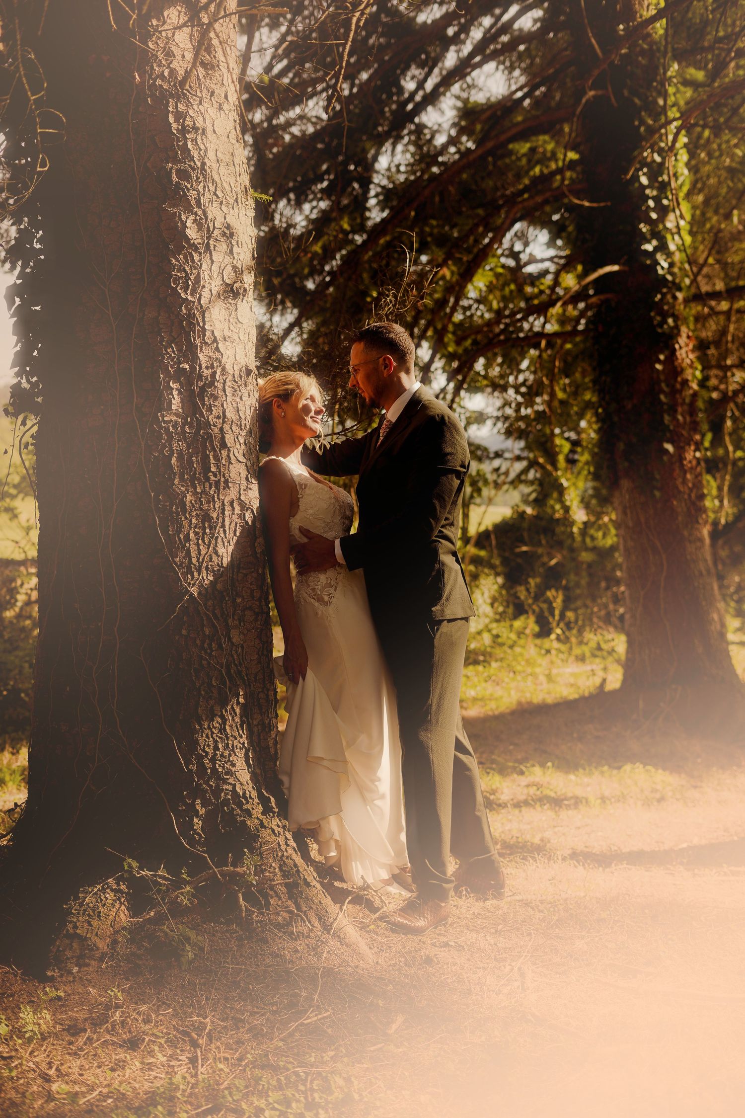 Bride and groom sharing intimate moment between sunlit trees in golden hour light.