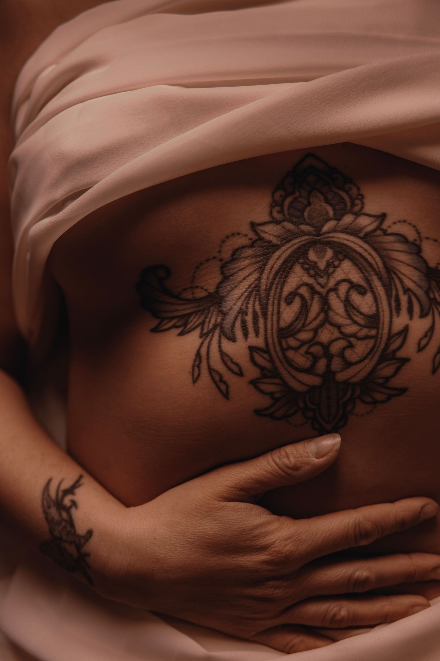 Close-up of an intricate ornate black tattoo on a torso wrapped in soft pink fabric with gentle hands.